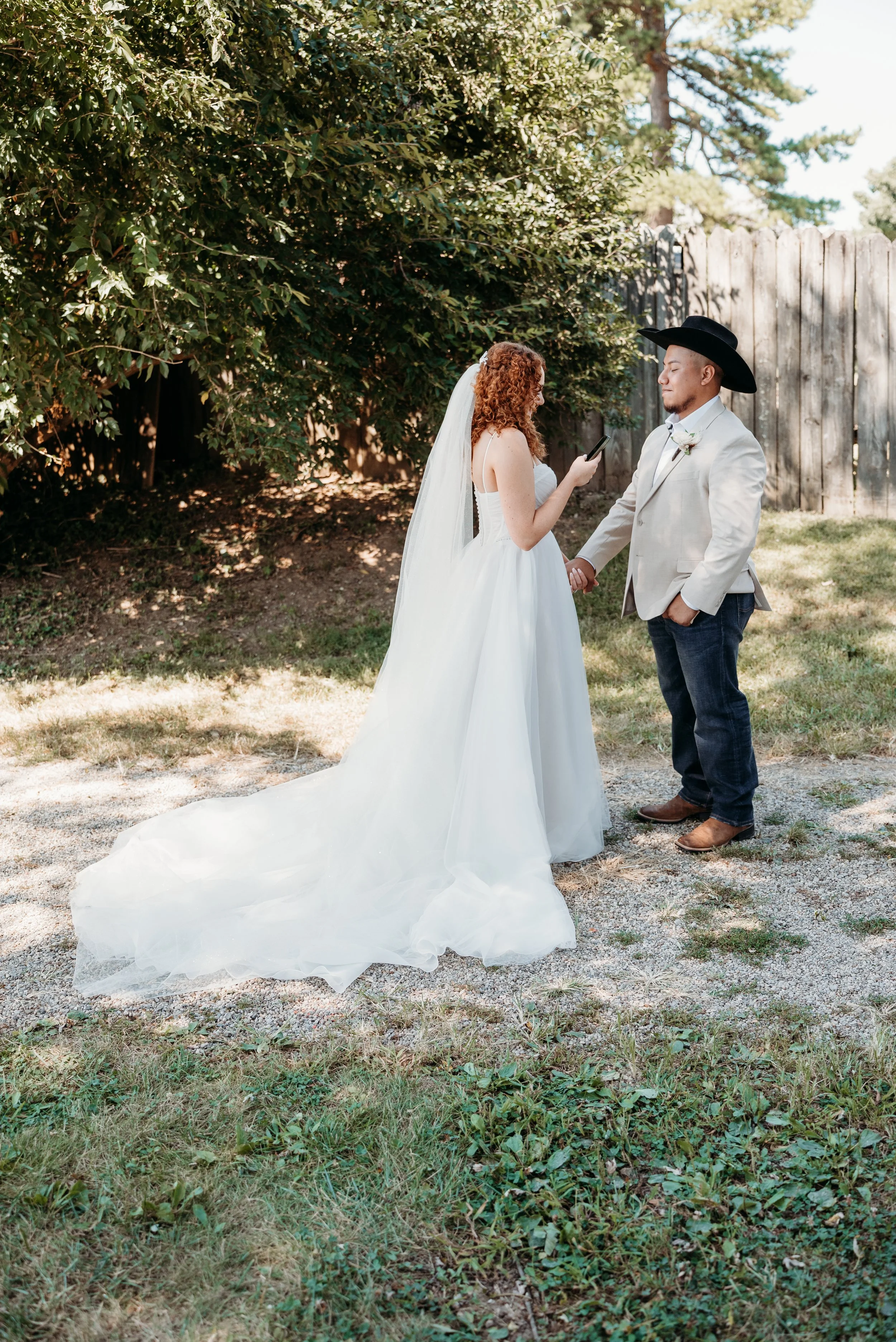 A bride and groom exchange vows outdoors, with the bride holding a phone, standing in front of greenery and wooden fencing.