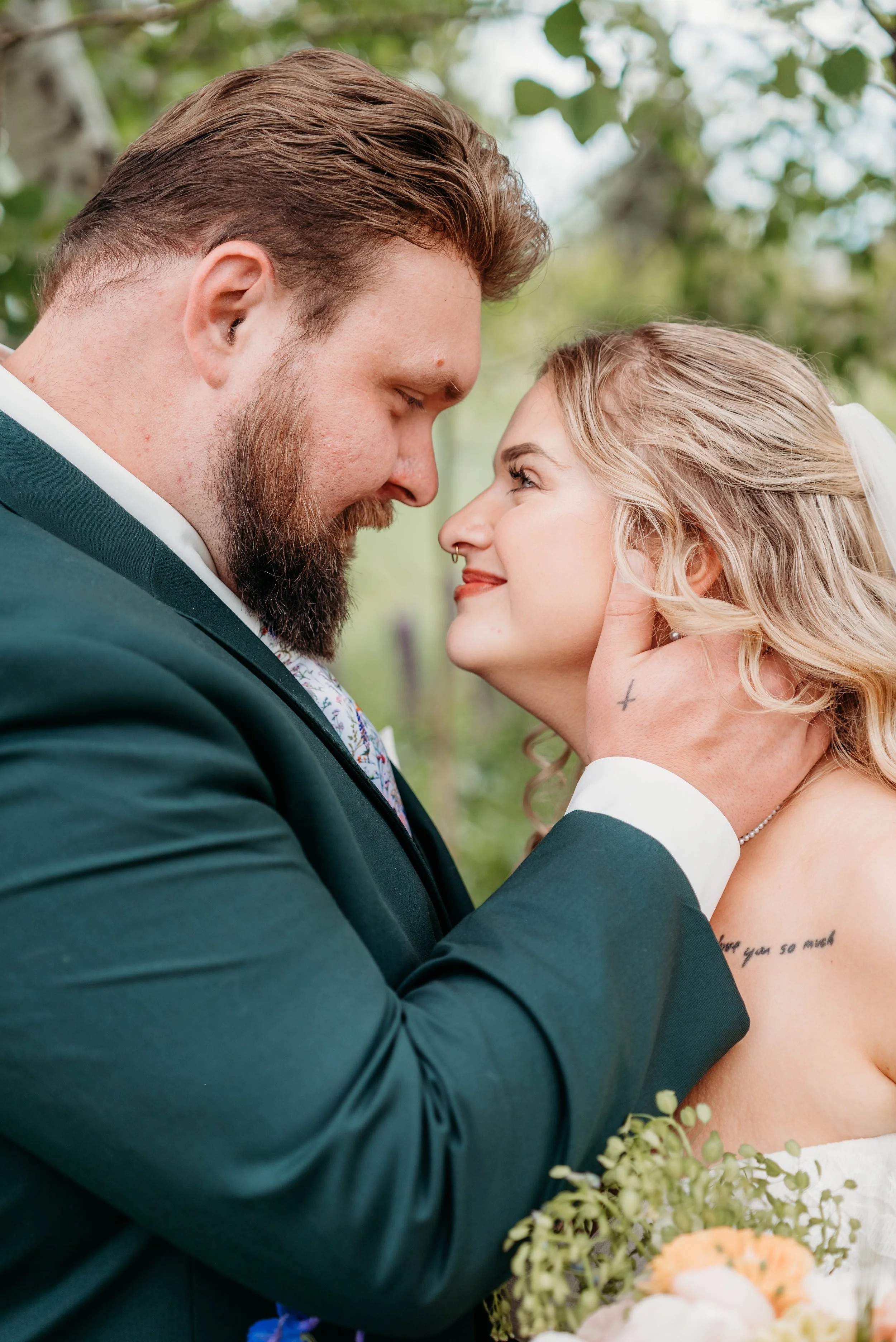 A bride and groom smiling closely, with their foreheads touching, outdoors with greenery in the background.