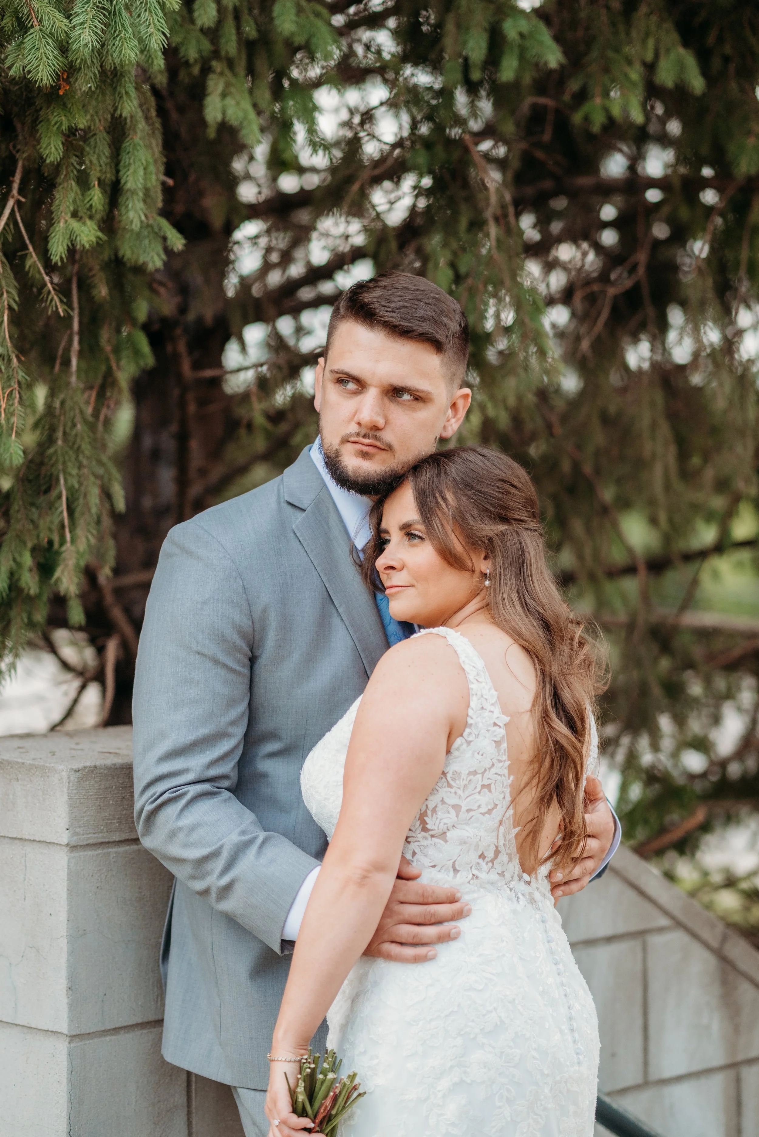 Bride and groom embrace outdoors, with lush green tree in background.