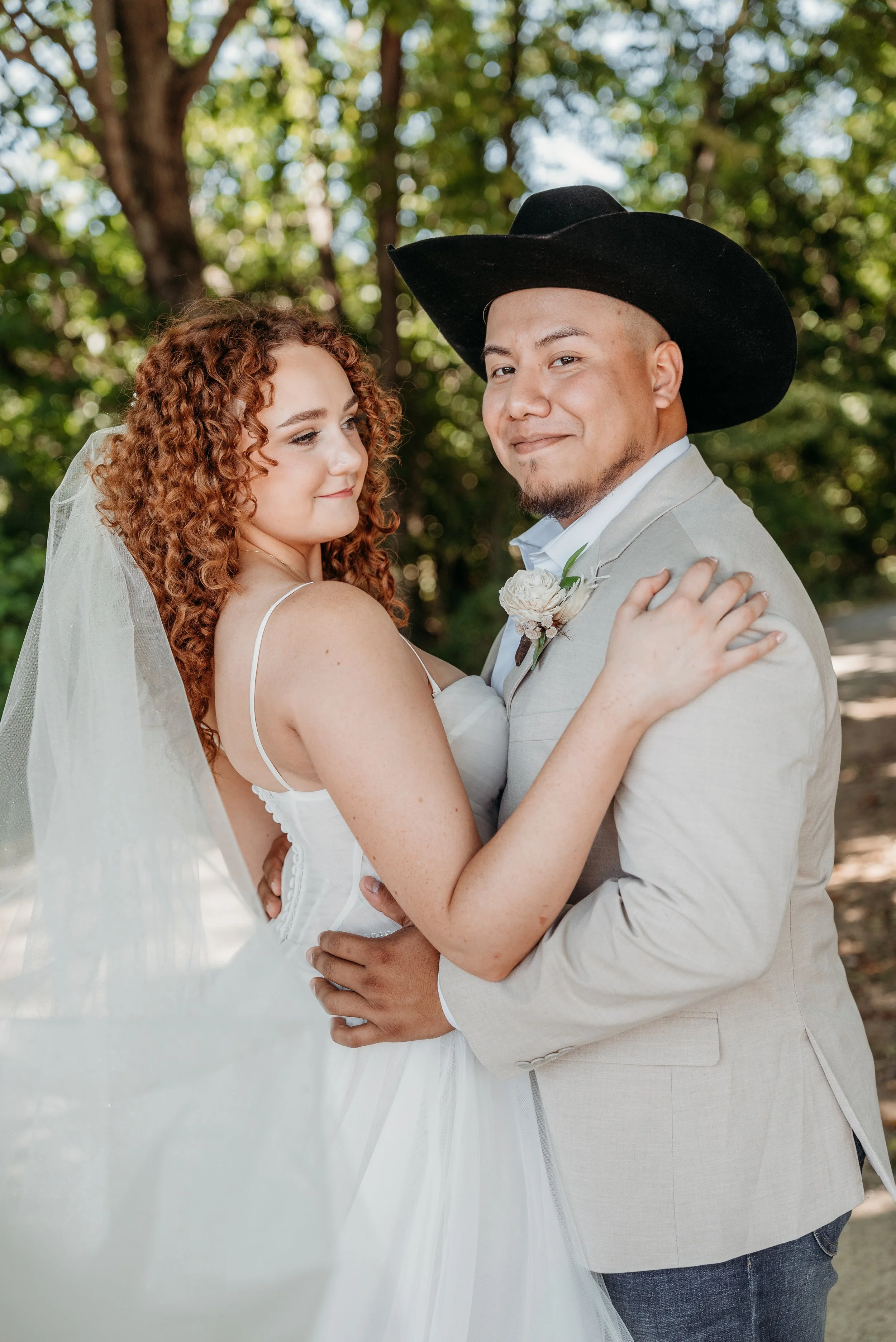 A bride with curly red hair and a groom wearing a cowboy hat, embracing each other outdoors surrounded by trees.