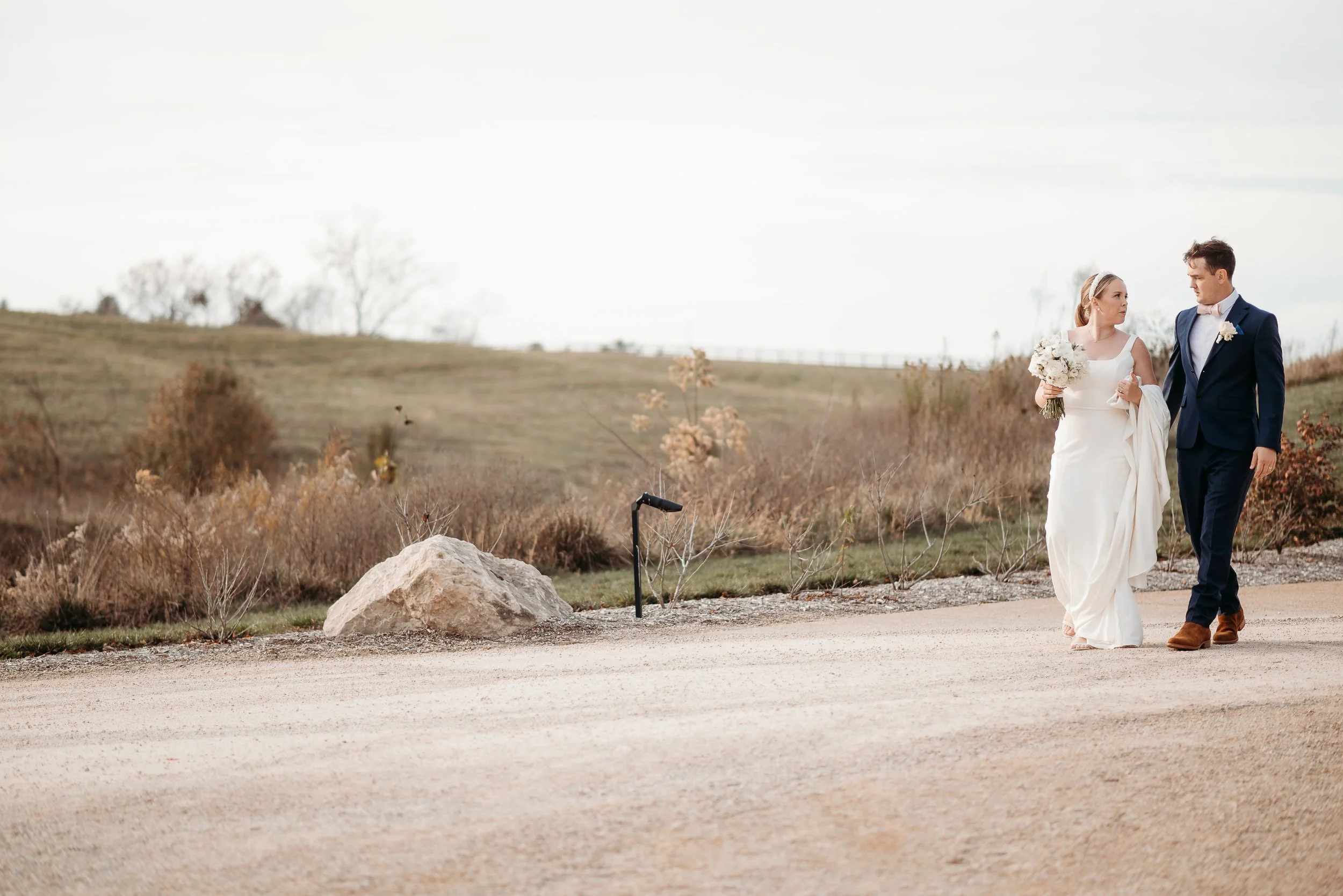 A bride and groom walking outdoors on a pathway, holding hands, with the bride holding a bouquet of white flowers, and the groom dressed in a navy blue suit. The landscape is open with a grassy field and some trees in the distance, under a cloudy sky.