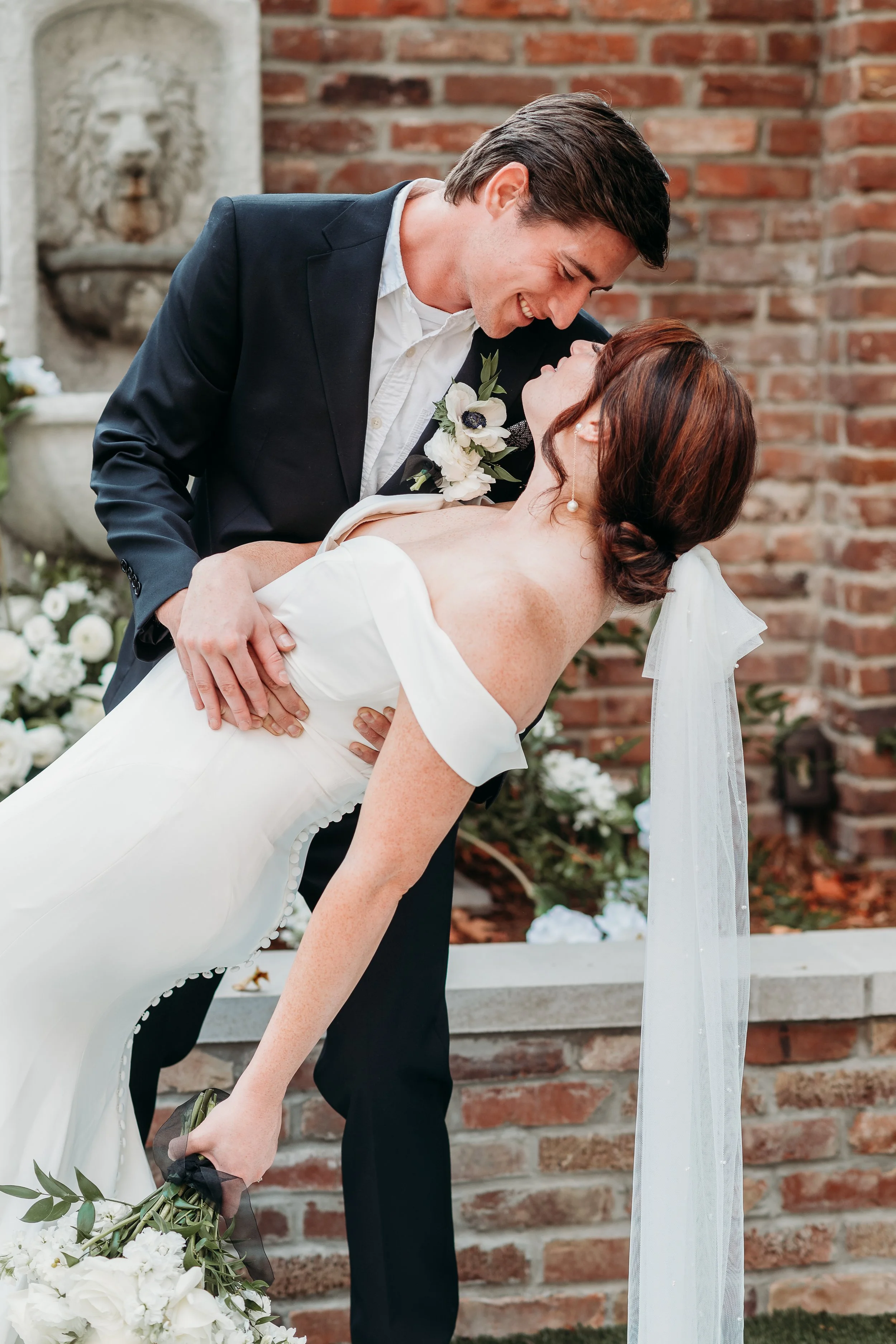 A couple shares a dance at their wedding, with the groom leaning in to kiss the bride who is dipping backward. The bride is in a white gown with off-the-shoulder sleeves and a veil, while the groom is in a dark suit with a white shirt and boutonniere. The background features a brick wall and decorative fountain.