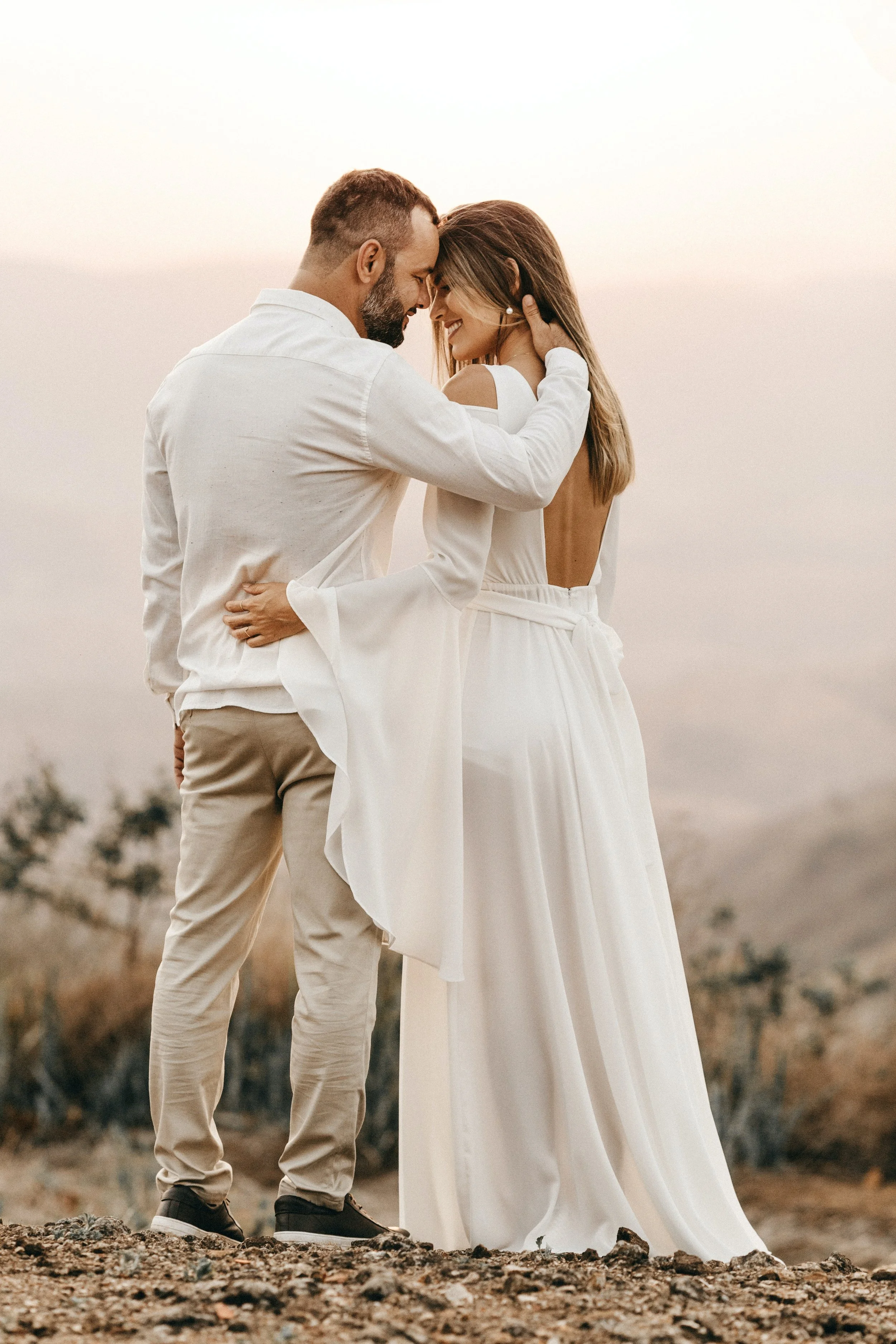 A couple dressed in white, with the man and woman sharing a tender moment outdoors, their foreheads touching, against a scenic, softly lit background.