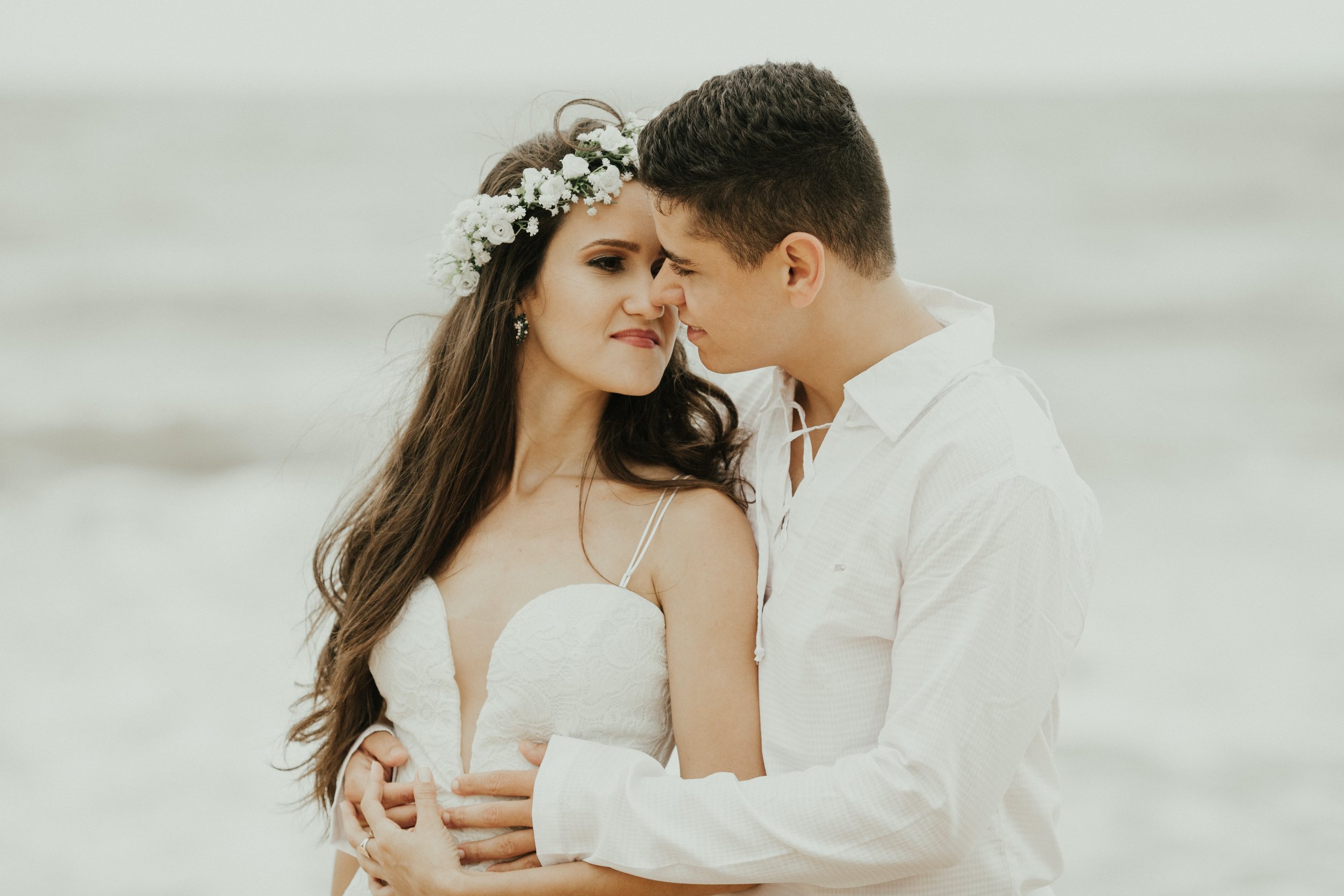 A couple in wedding attire close together on a beach, touching foreheads, with a calm, light-colored background.