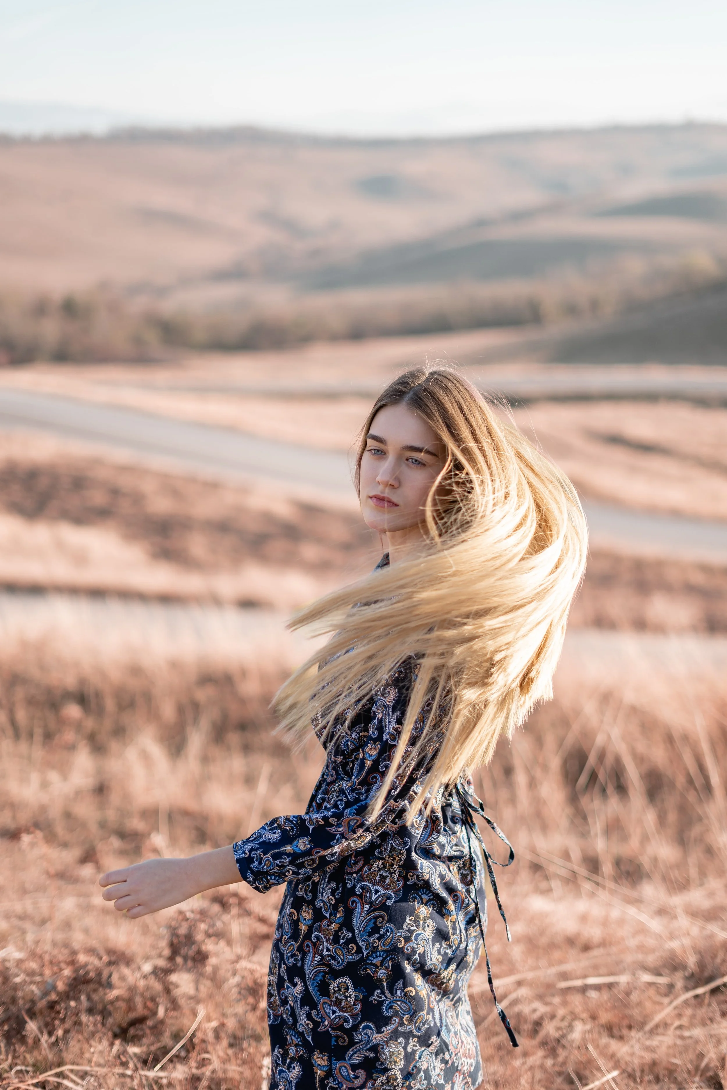 A young woman with long blonde hair standing in a dry, hilly field with a blurred landscape of rolling hills in the background.
