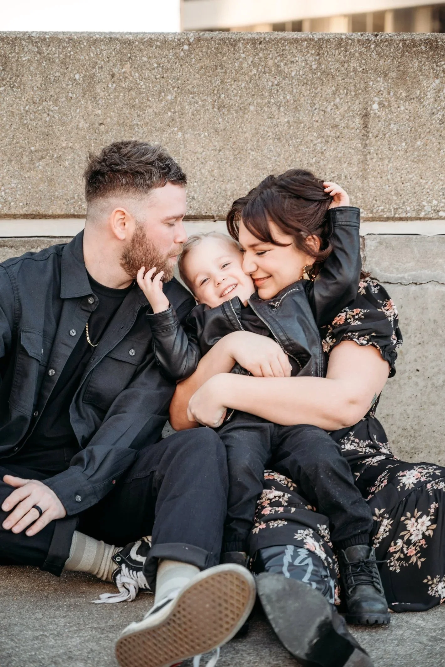 A family of three, a man, a woman, and a child, sitting on the ground against a concrete wall, embracing and smiling at each other.