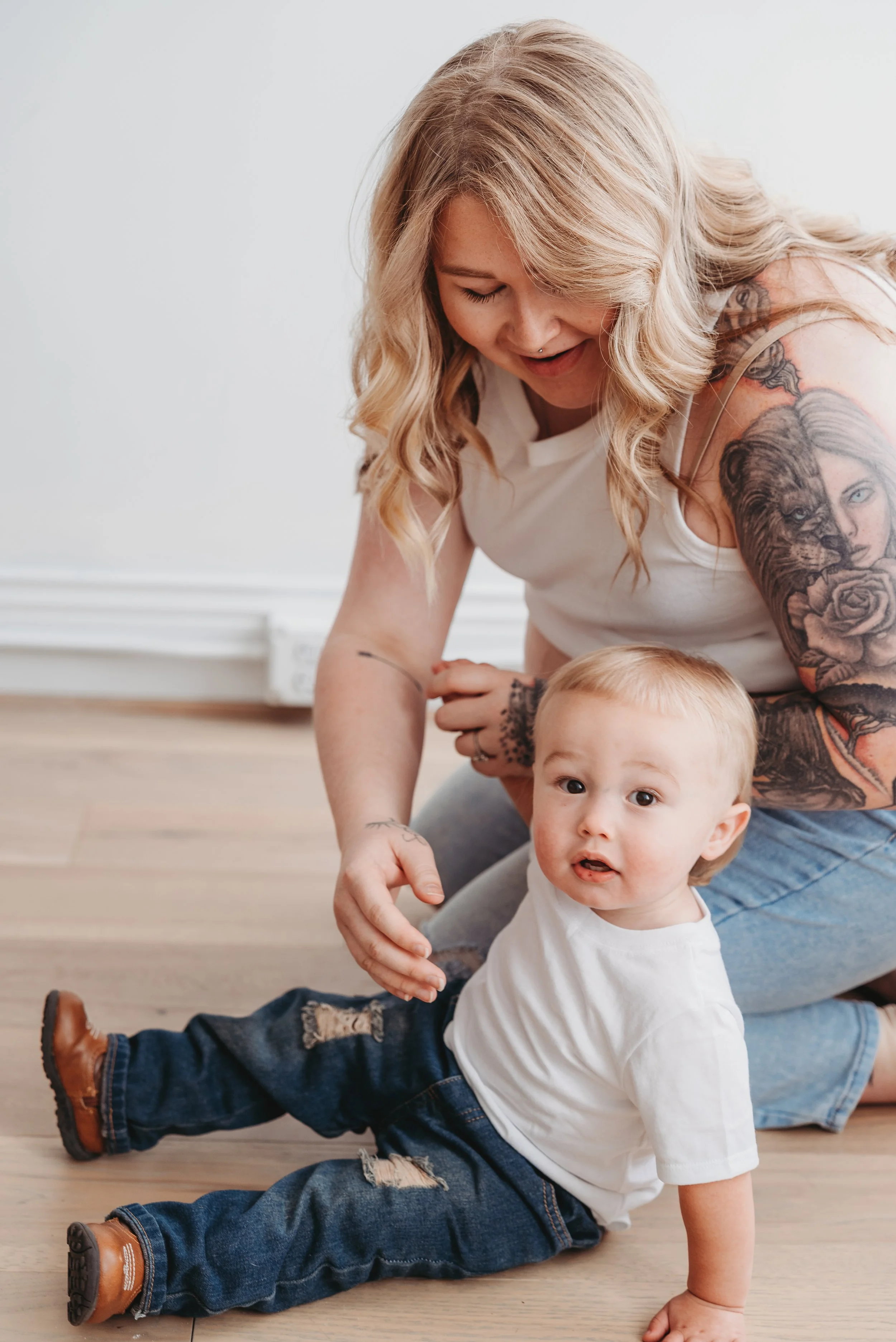 A woman with tattoos on her arms sitting on the floor with a young boy, who is sitting on the floor, looking at the camera, next to a white wall.