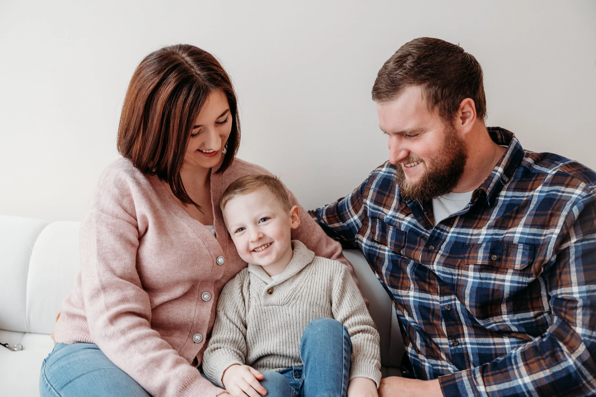 A smiling family of three sitting on a white couch, with a woman and man looking at their young son who is sitting between them, all happily engaged.