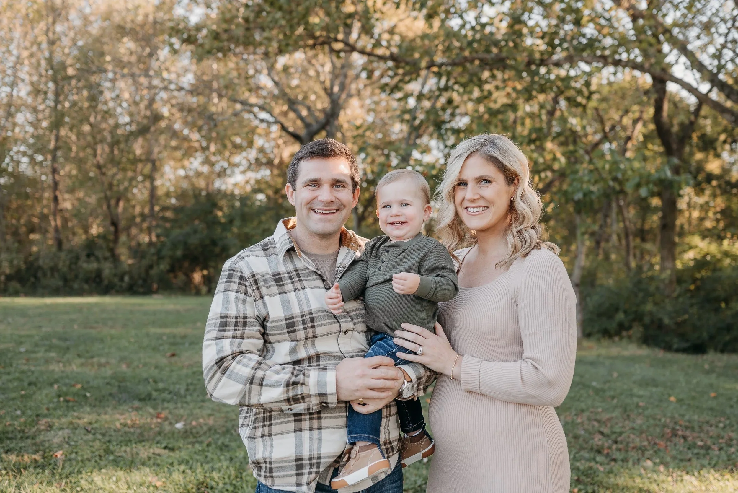 A family of three smiling outdoors during daytime, with a man, woman, and a young child, standing in a grassy area with trees in the background.