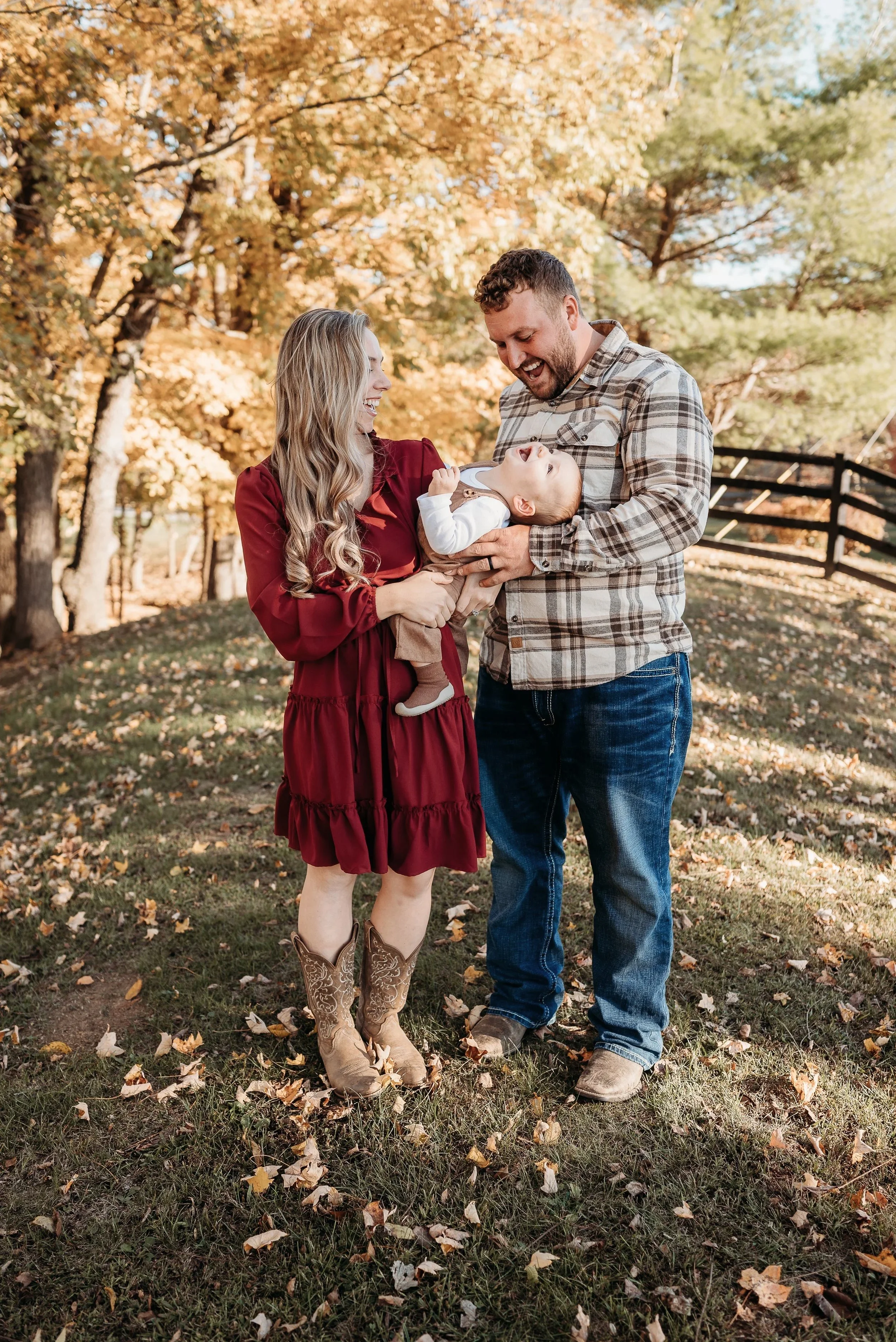 A family of three, including a woman in a red dress and cowboy boots, a man in a plaid shirt and jeans, and a baby, enjoying a sunny autumn day outdoors surrounded by trees with fall foliage and fallen leaves on the ground.