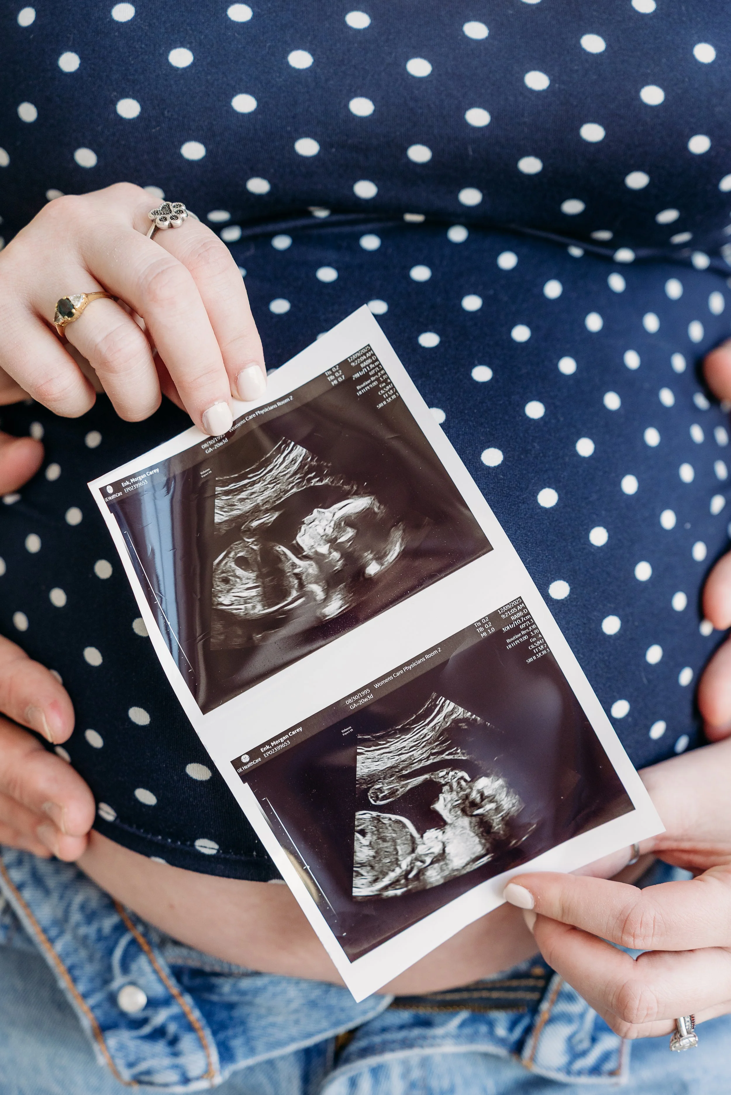 Person holding two ultrasound images over their pregnant belly, wearing a navy blue polka dot top and denim jeans.