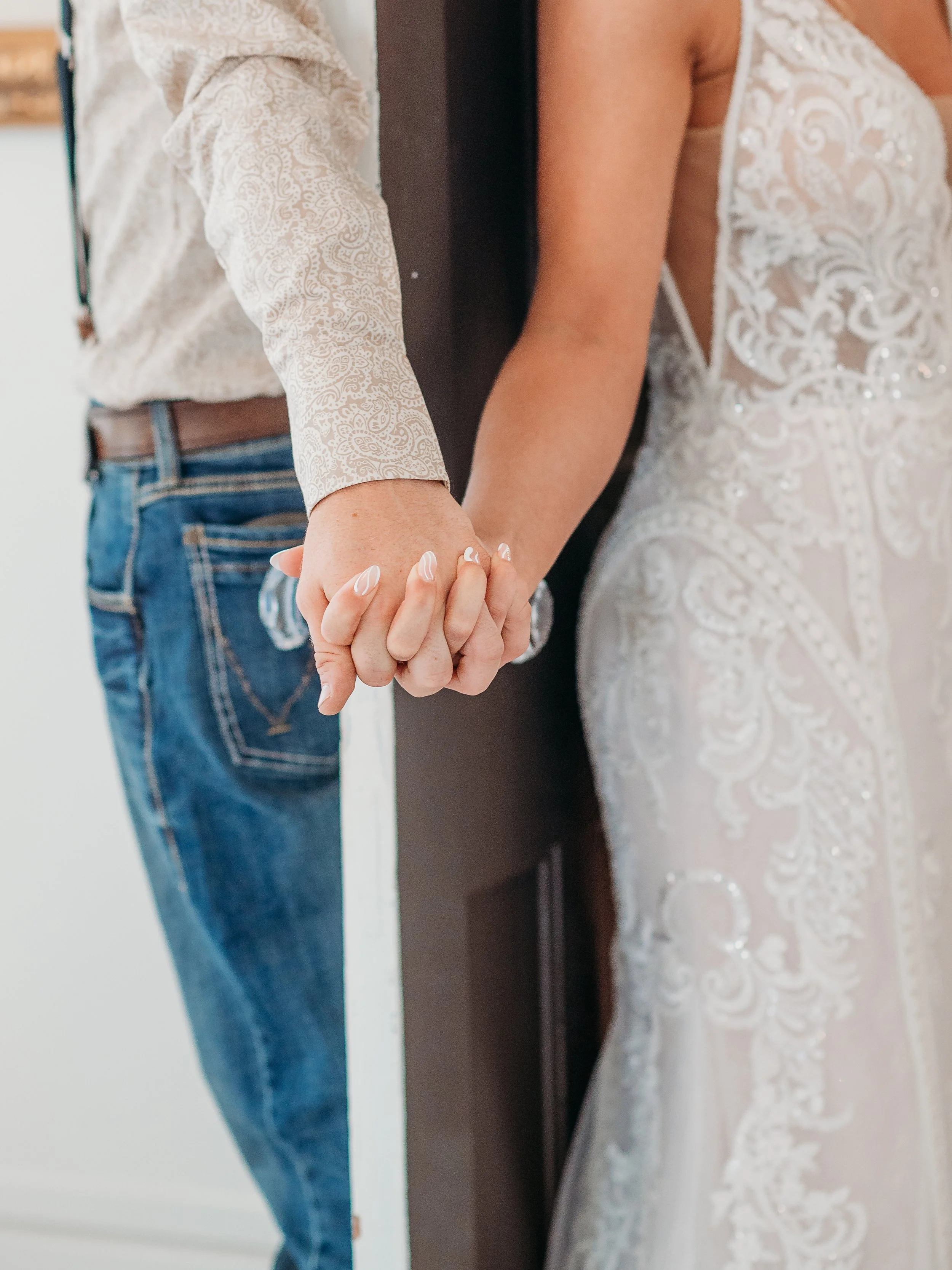 A couple holding hands with wedding attire, the woman in a lacy wedding dress and the man in a patterned shirt and jeans.