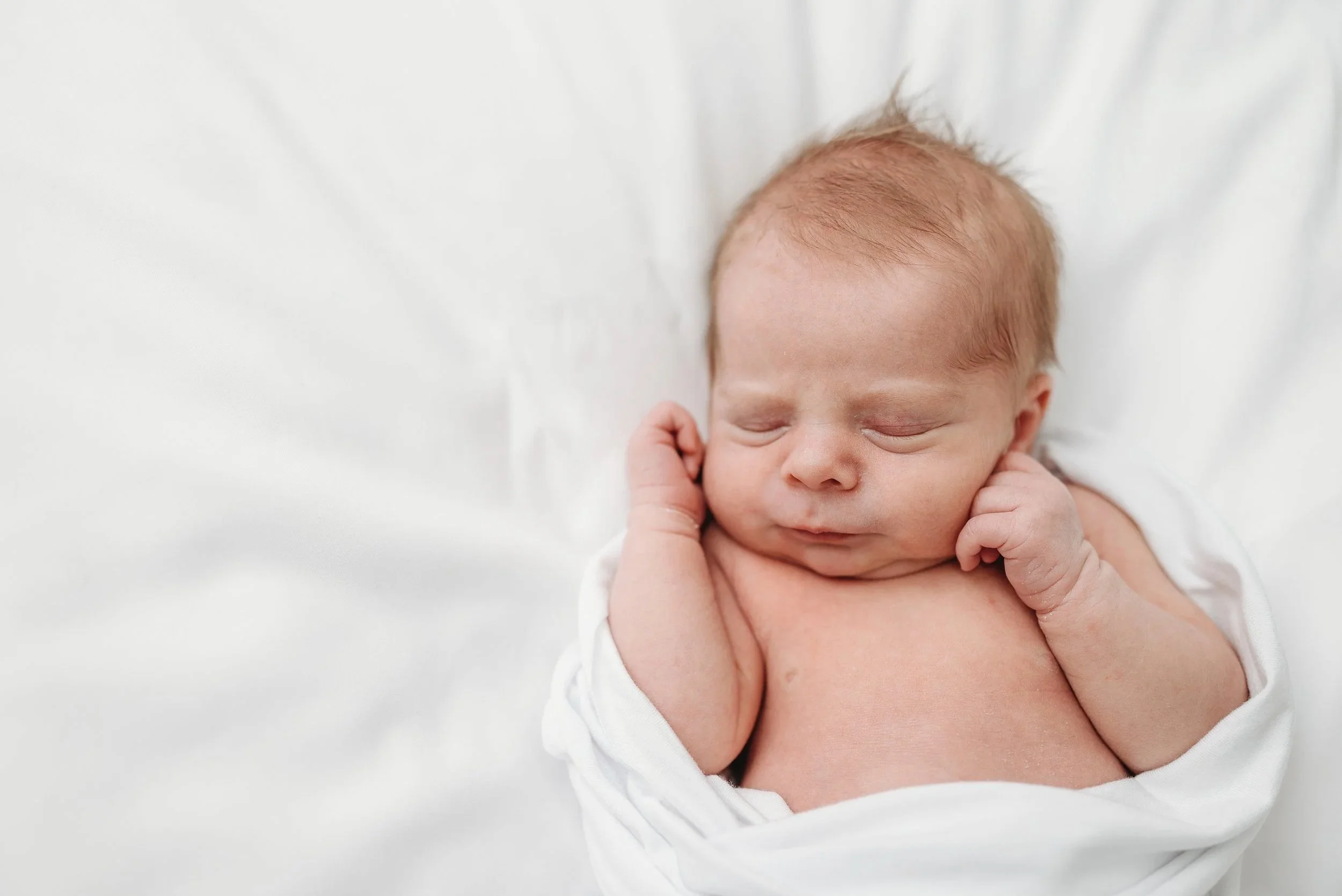 Newborn baby with red hair sleeping on a white blanket, swaddled in a white cloth.