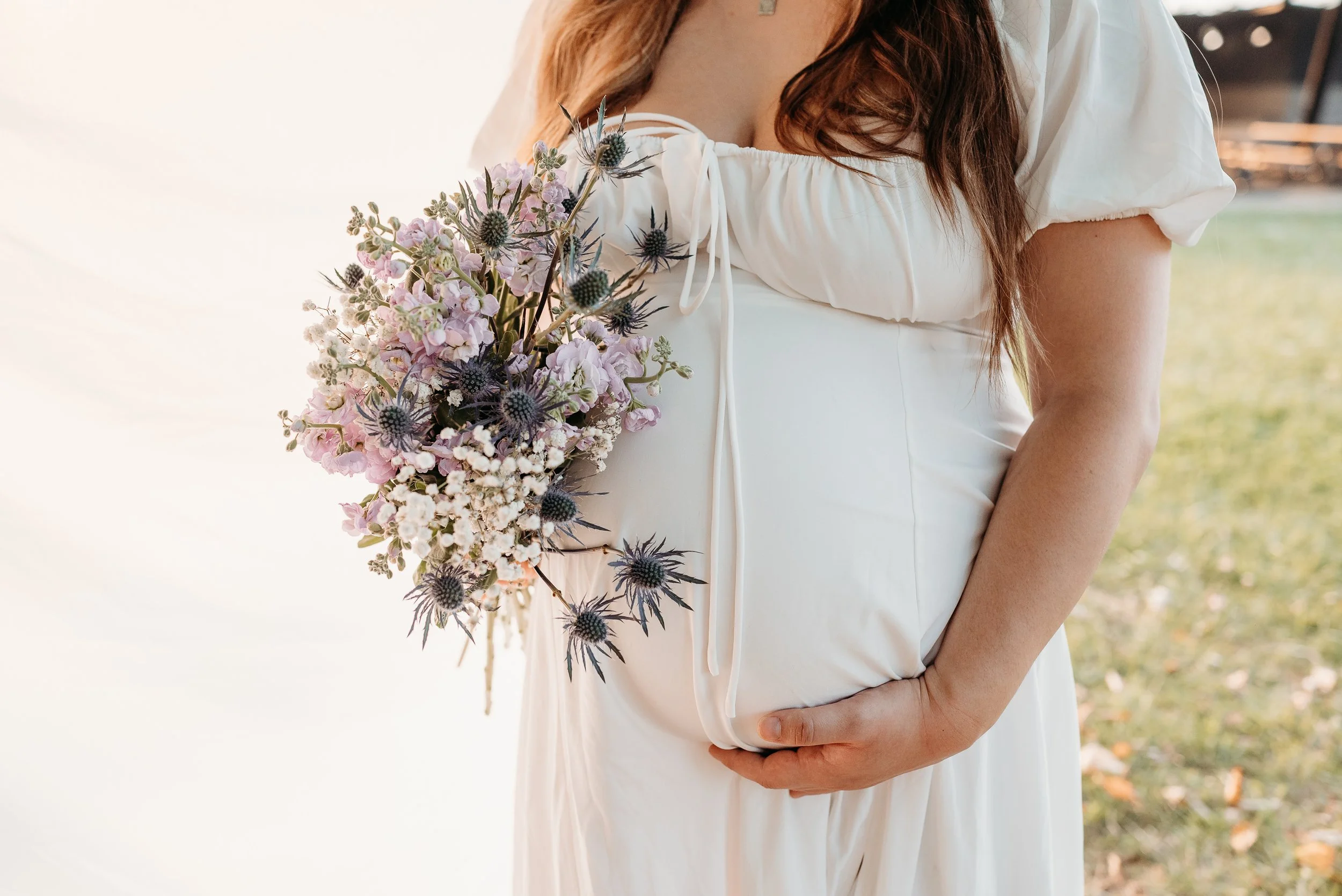 A woman in a white dress holding a bouquet of pink and purple flowers, standing outdoors.