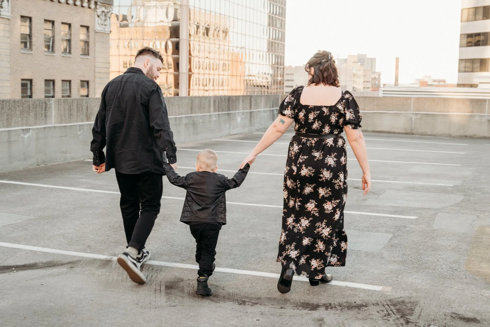 A family walks hand in hand in an empty parking lot with a cityscape in the background, during sunset.