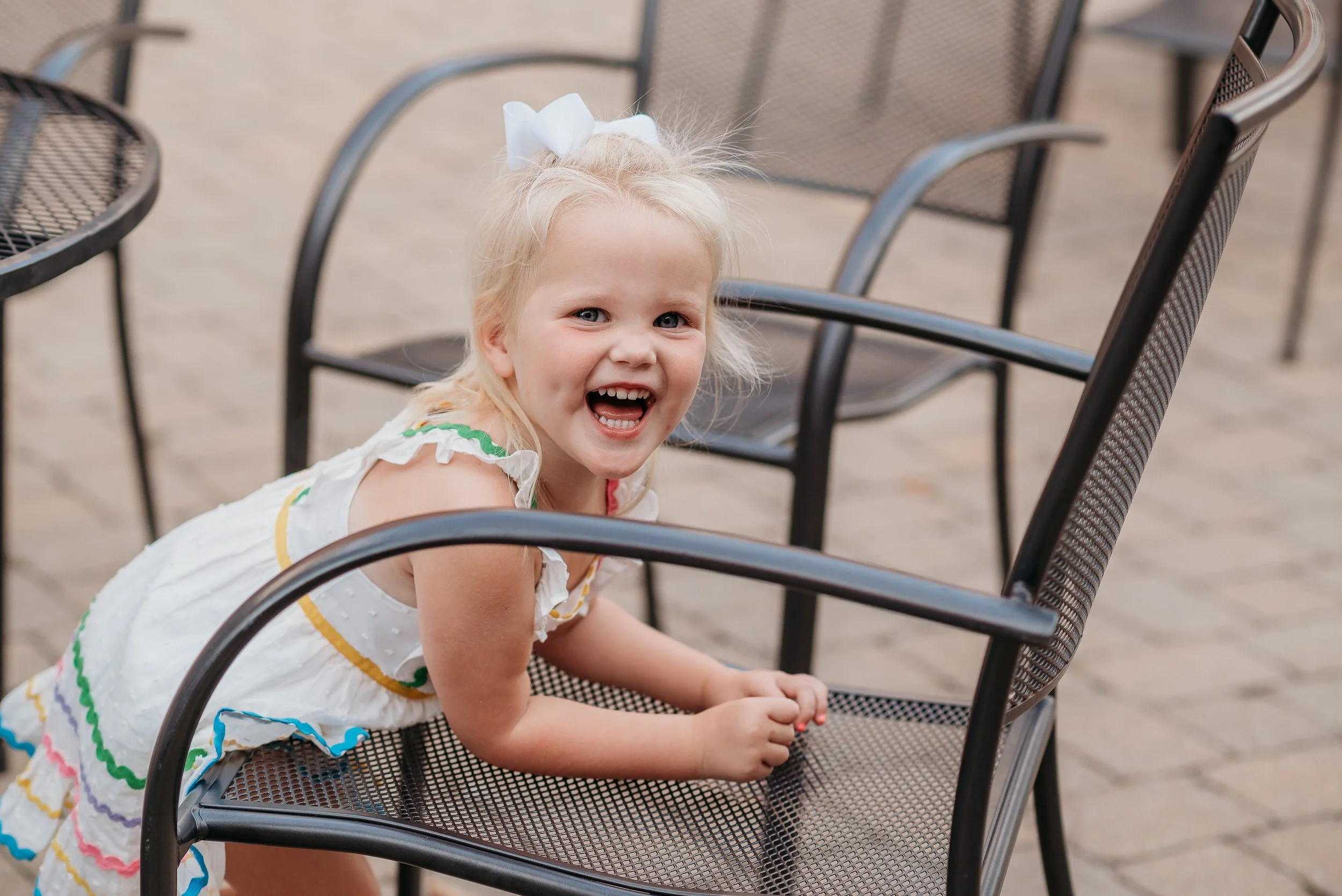 A young girl with blonde hair and a white bow, wearing a white dress with colorful trim, leaning on a black metal chair, smiling and looking at the camera.