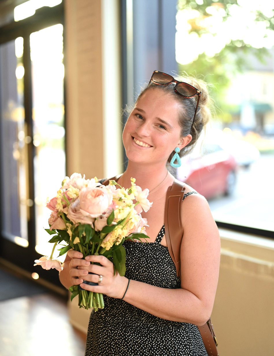 A young woman with sunglasses on her head, earrings, and a black dress with white polka dots, holding a bouquet of light pink and white flowers, smiling indoors near a window.