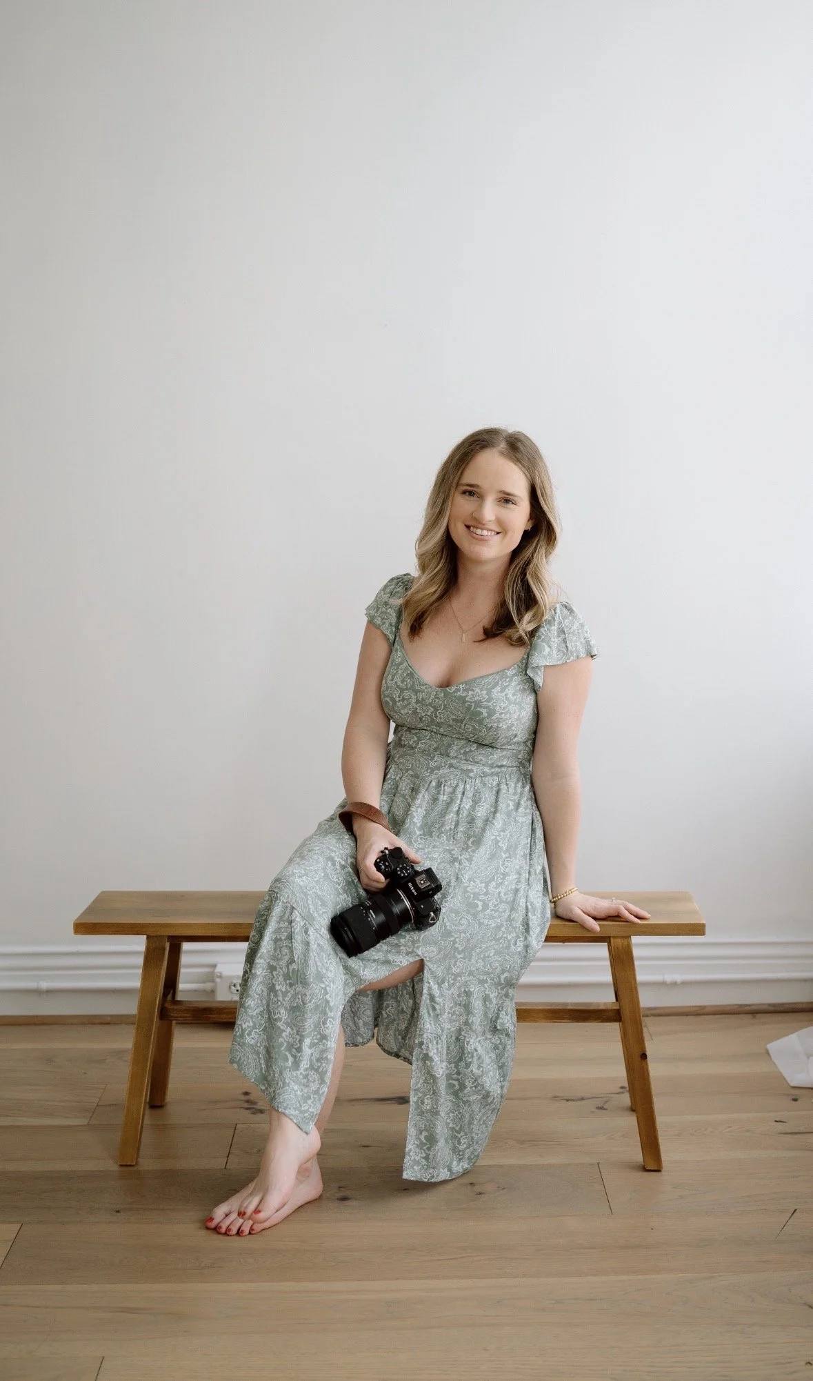 A woman with blonde hair, dressed in a green patterned dress, sitting barefoot on a wooden bench in a room with a white wall and wooden floor. She is holding a black camera with a lens in her right hand, smiling at the camera.