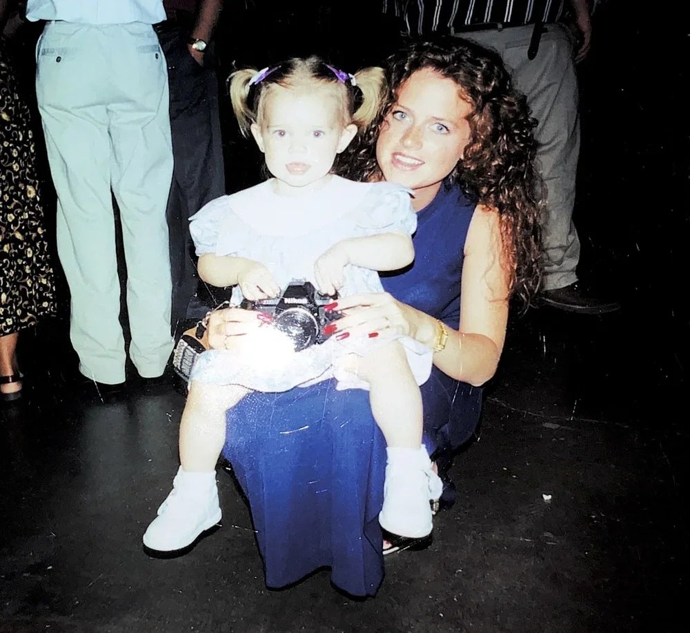 Woman with curly hair and blue dress holding young girl with pigtails and white dress, at a social event.