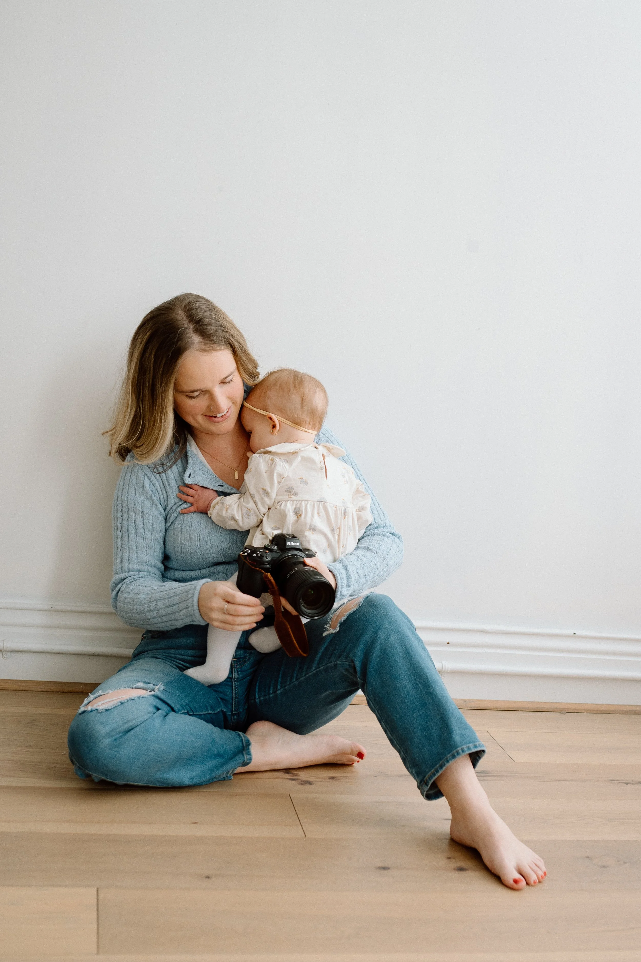 A woman sitting on the floor holding a camera in her lap, with a young child on her lap, both smiling and leaning against a white wall.