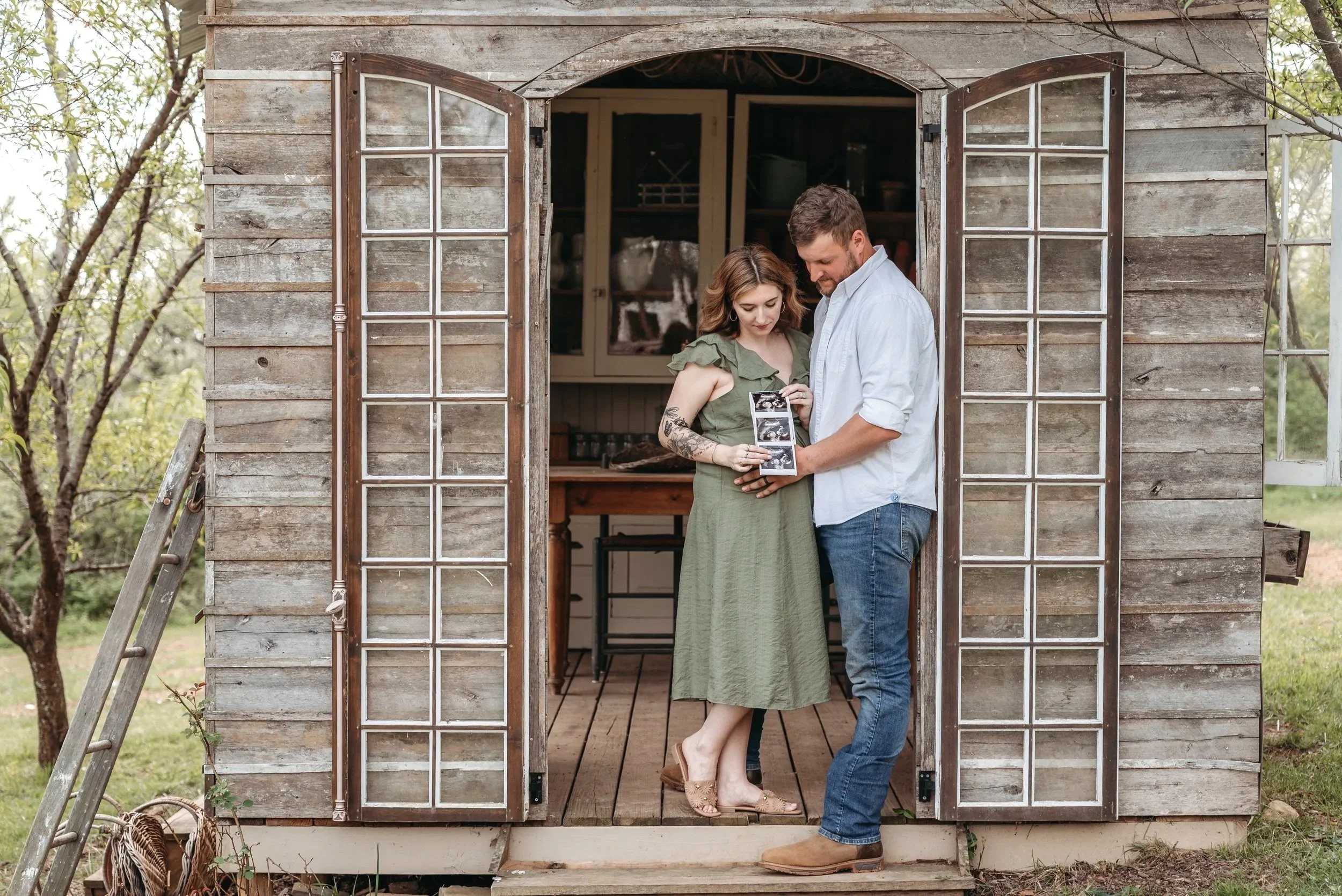 A couple standing in the doorway of a rustic wooden shed, looking at ultrasound pictures together, surrounded by trees and outdoor greenery.