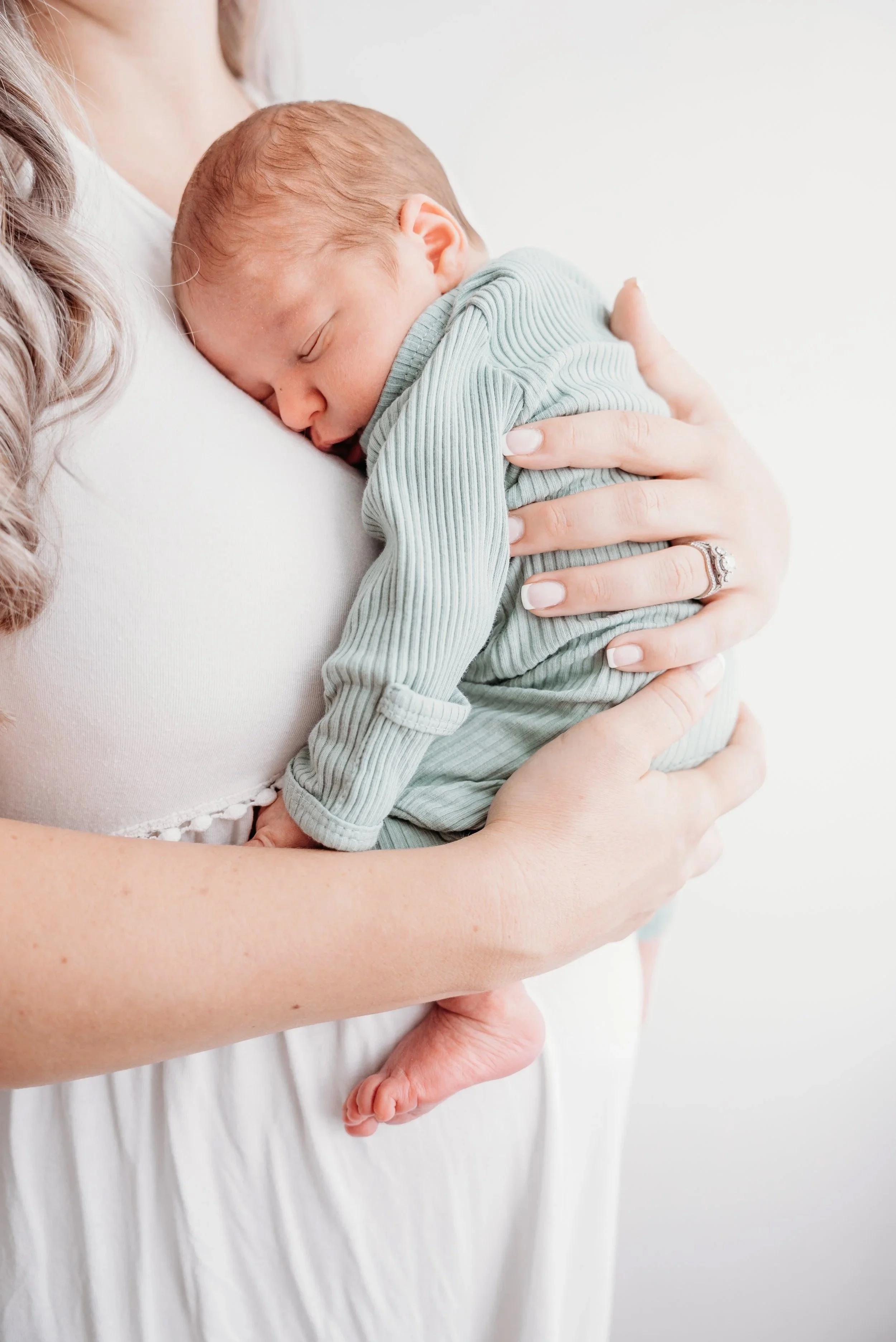 A woman holding a sleeping baby close to her chest, with her hand resting on the baby's back. The baby is wearing a green striped outfit and is resting peacefully. The background is plain white.