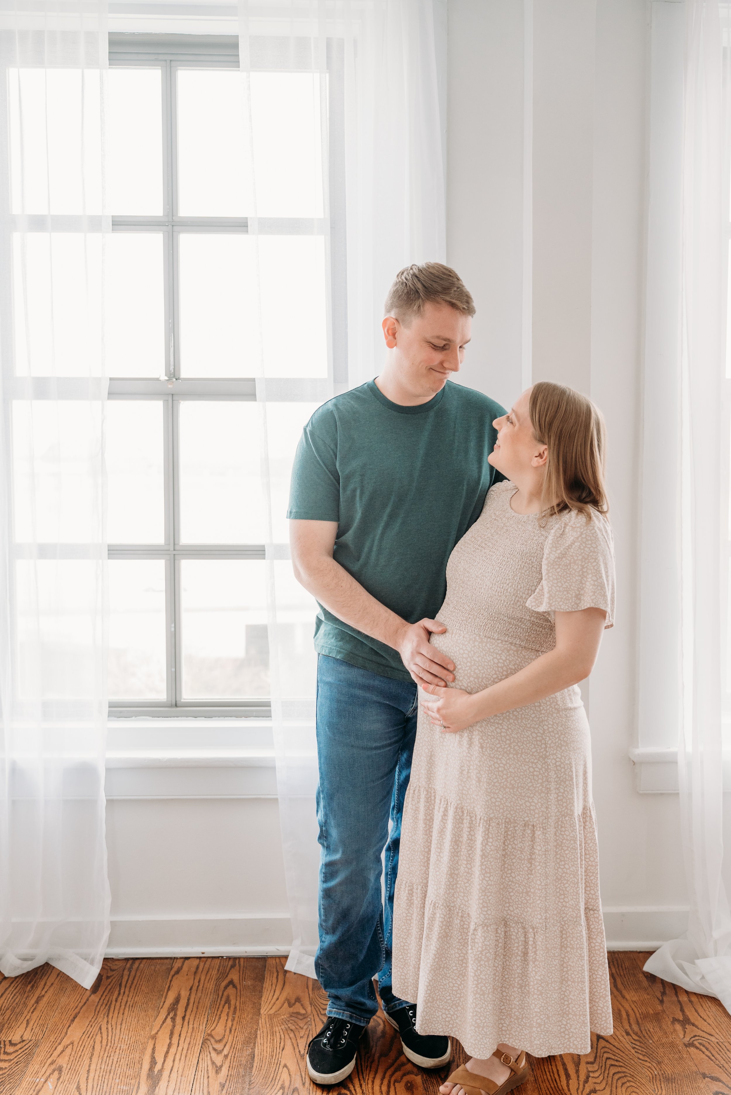 A man and woman standing close together in a bright room with large windows and white curtains, the man touching the woman's pregnant belly and both smiling softly at each other.