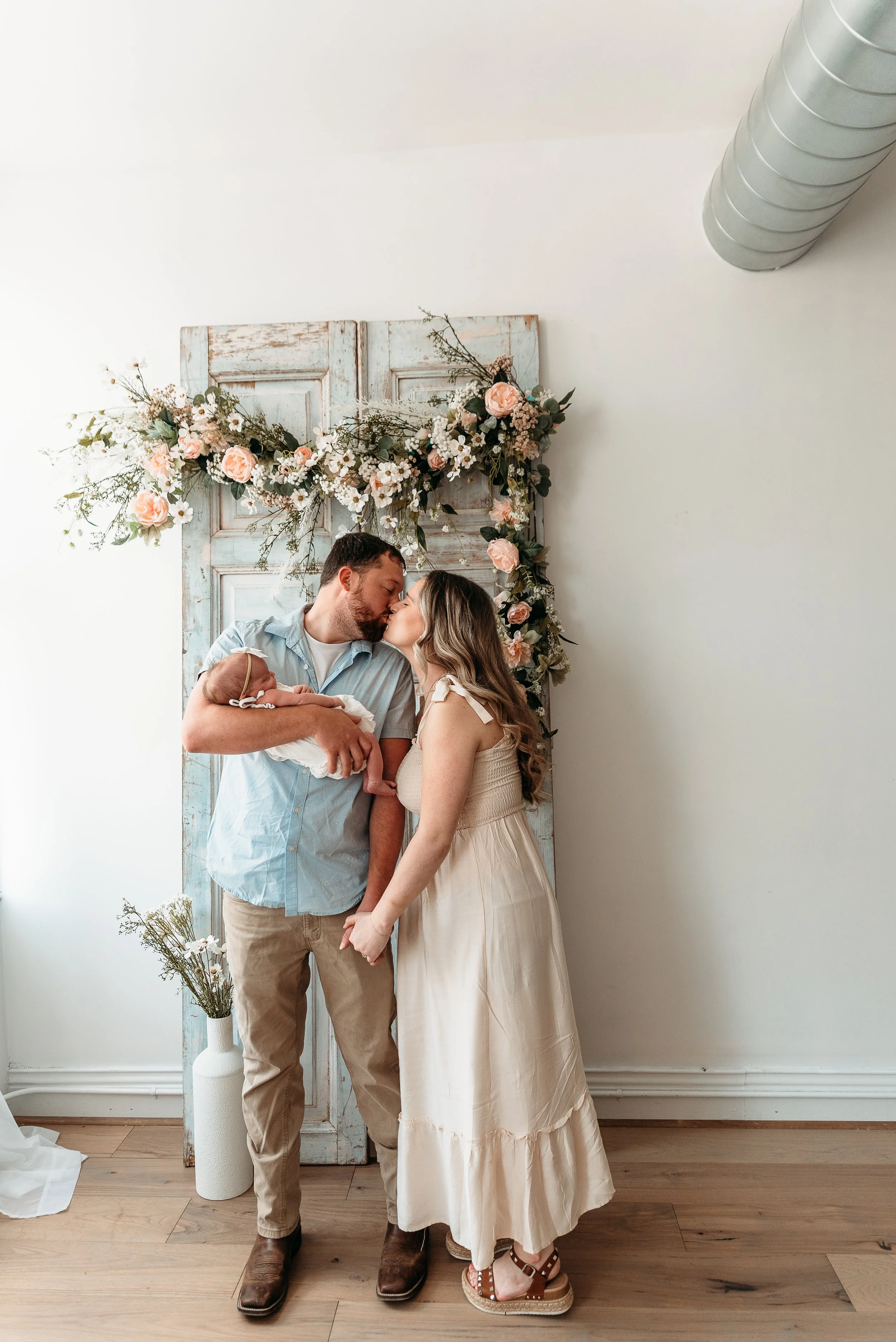A couple holding a newborn baby and kissing, standing in front of a rustic floral backdrop with pink roses and white flowers, in a bright room with wooden floors.