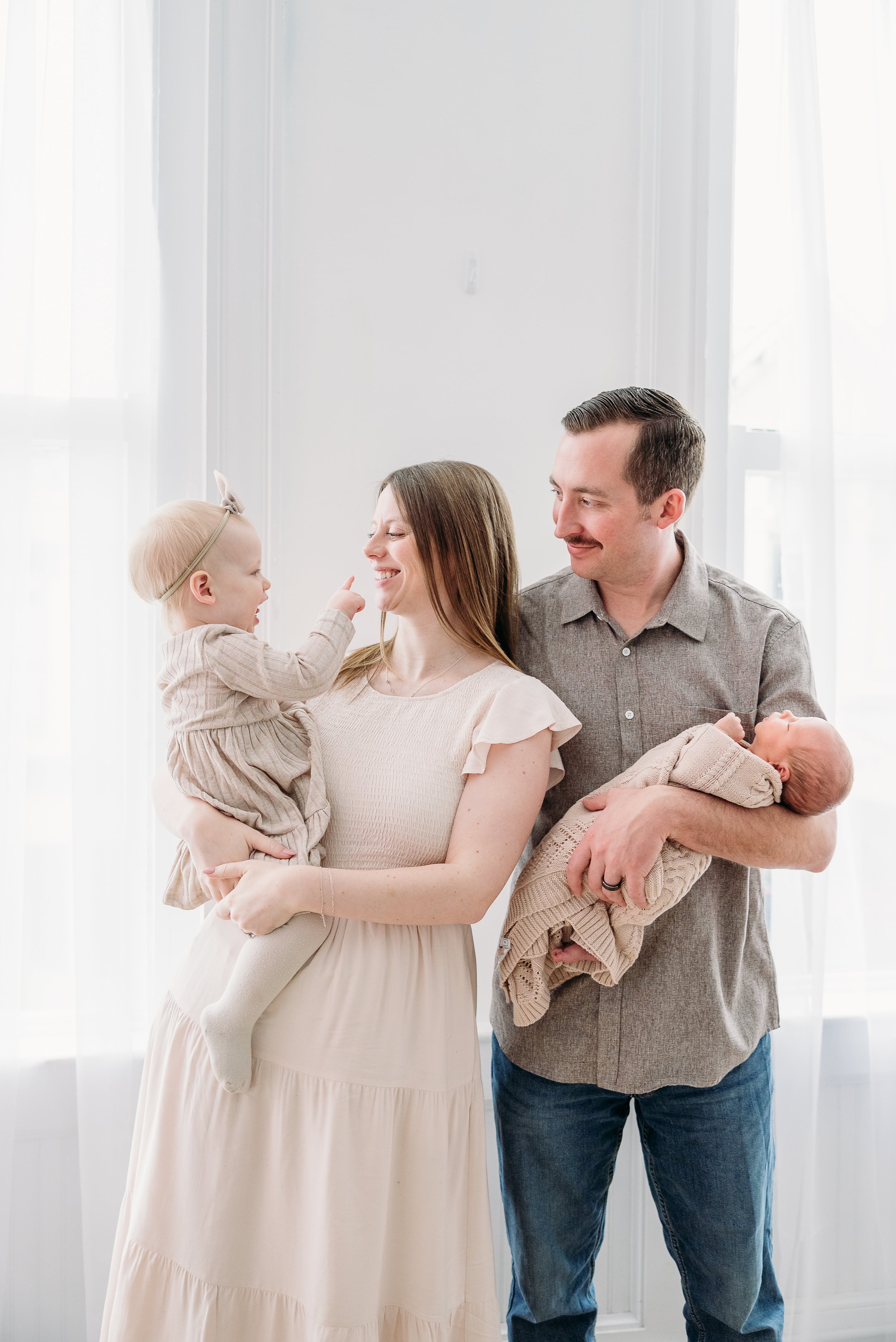 Family with young child and newborn baby in a bright room with white curtains.