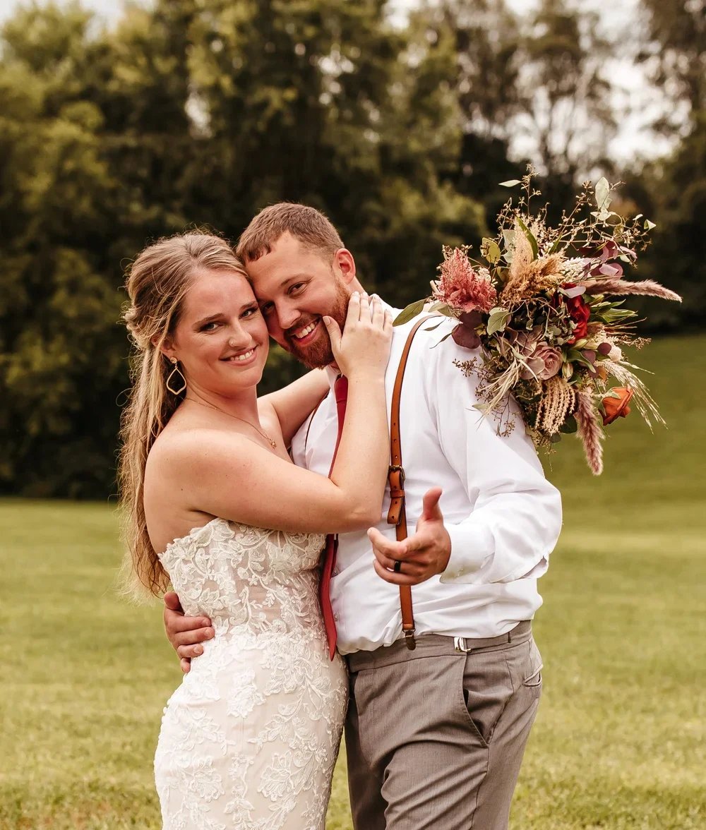 A smiling bride and groom embrace outdoors, the bride in a lace wedding dress and the groom with a bouquet of flowers on his shoulder, on a grassy field with trees in the background.