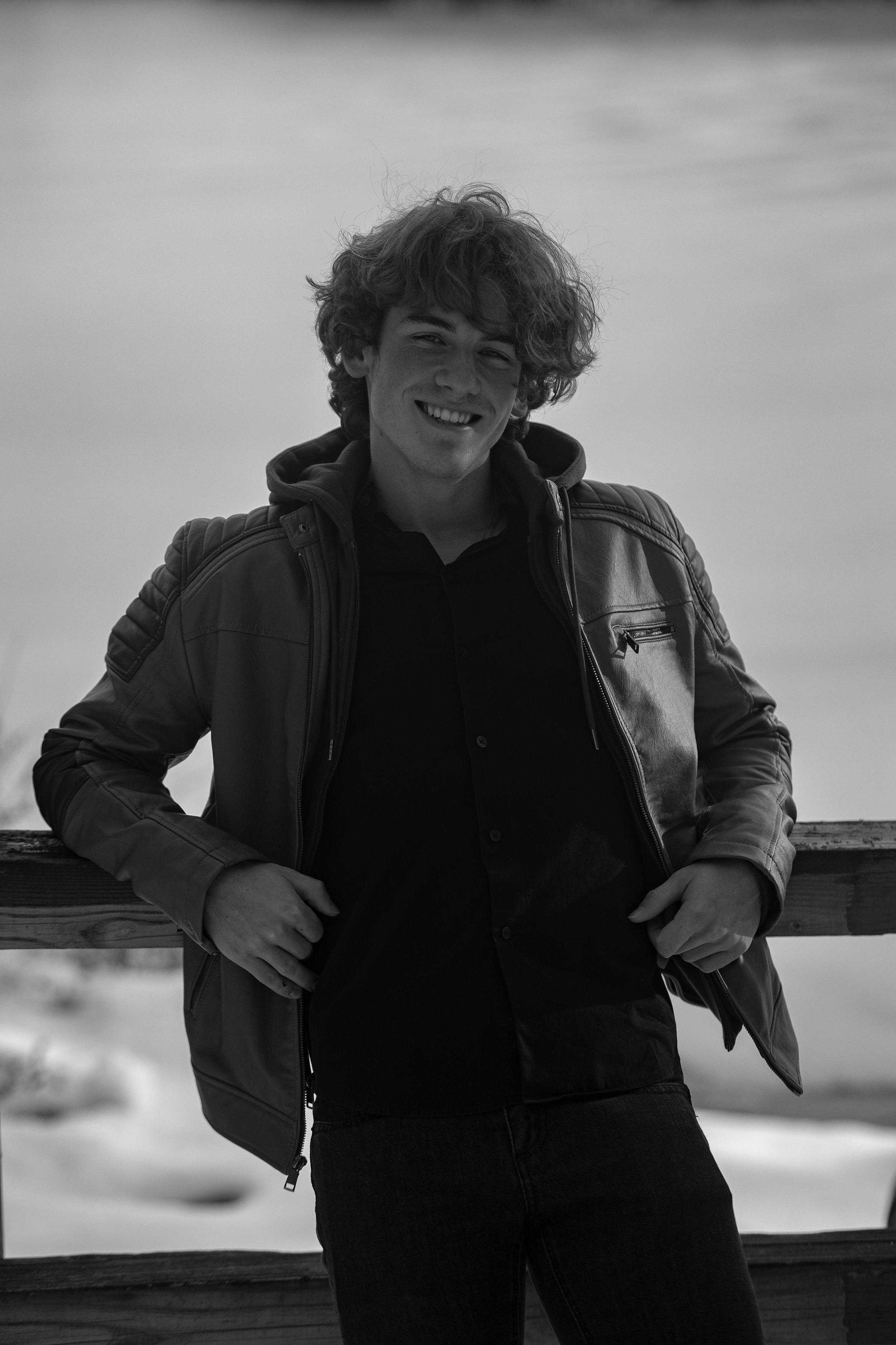 A young man with curly hair smiling and leaning on a wooden fence outdoors.