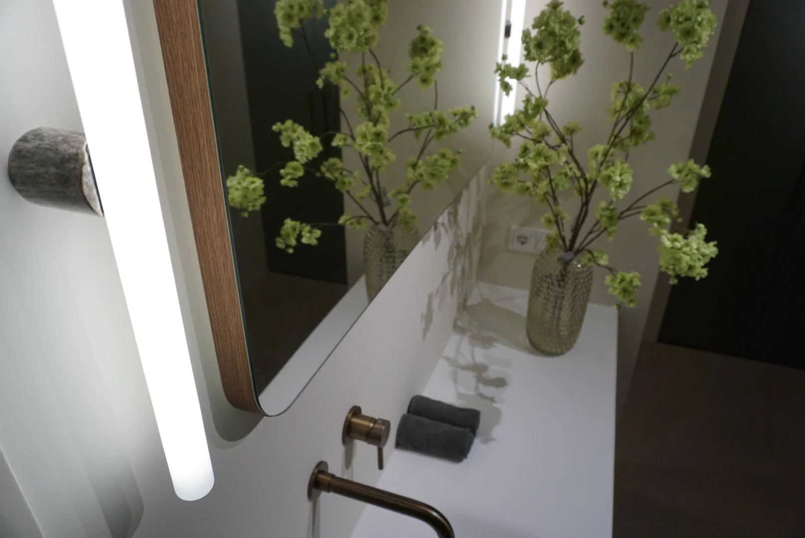 Een witte badkamer spiegel met een houten rand, naast twee vazen met groene bloemen op een wc-botten. Verder op de rand liggen een zwarte handdoek en twee koperen armaturen.