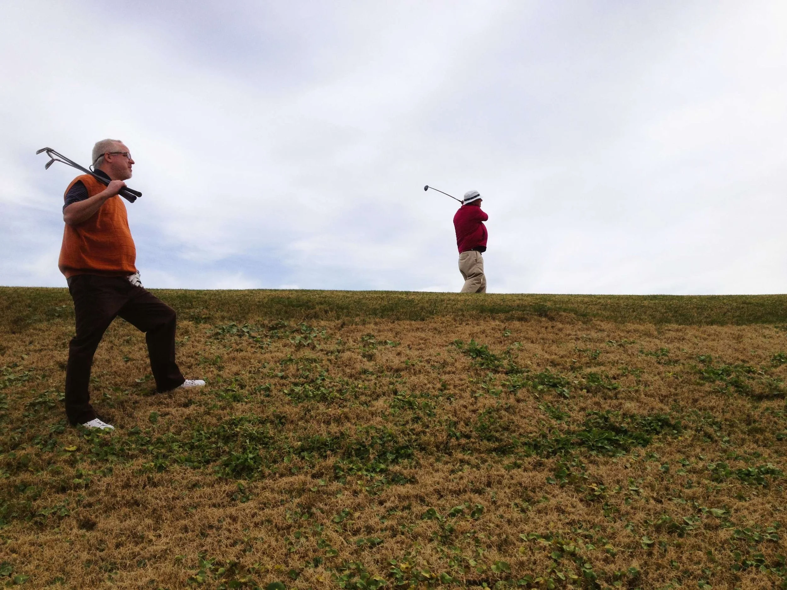 Two men playing golf on a grassy course, one walking with a club over his shoulder and the other preparing to swing a golf club.