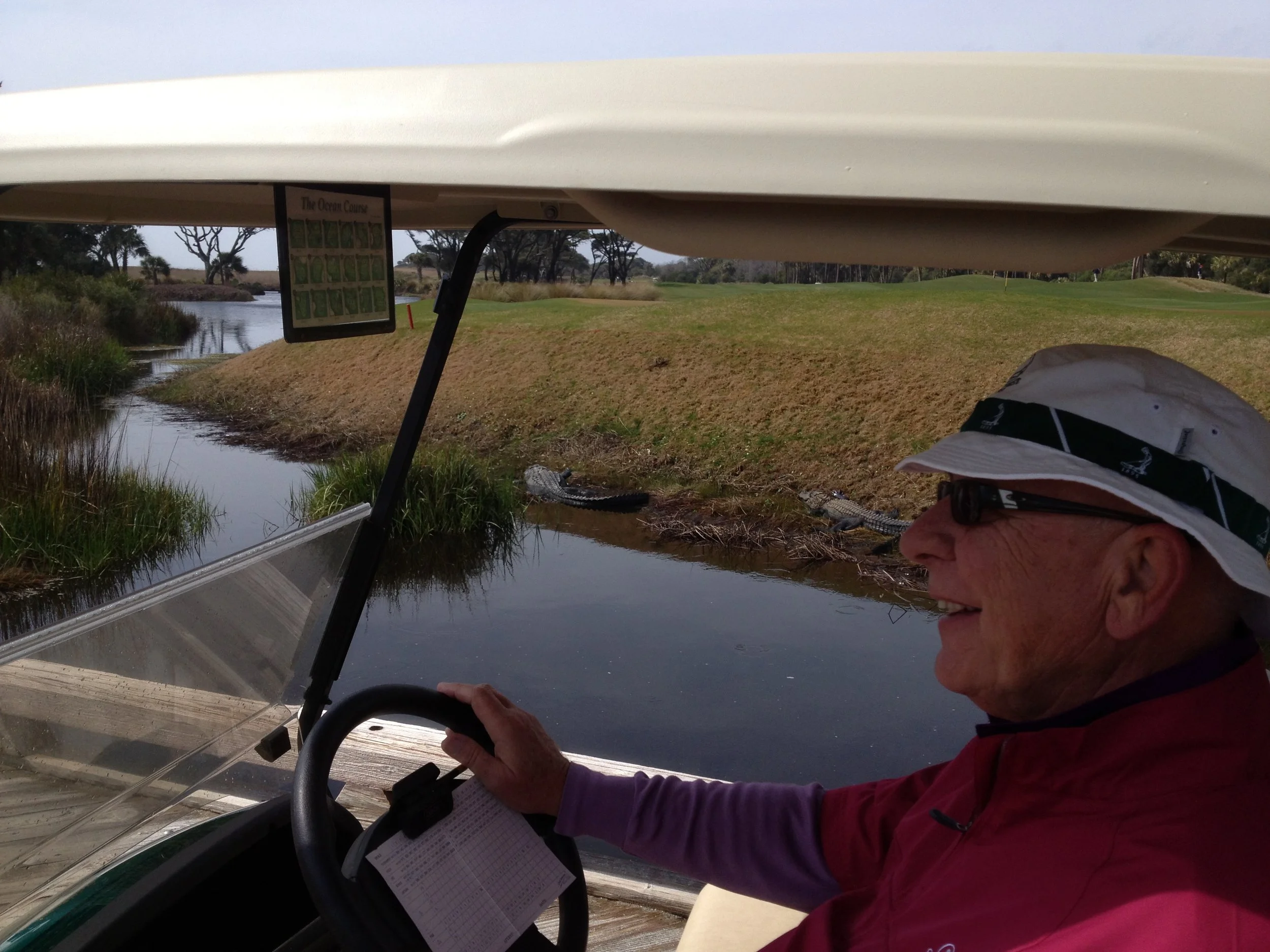 An older man smiling and driving a golf cart on a golf course, with a water hazard and greenery visible in the background, wearing a sun hat and glasses.