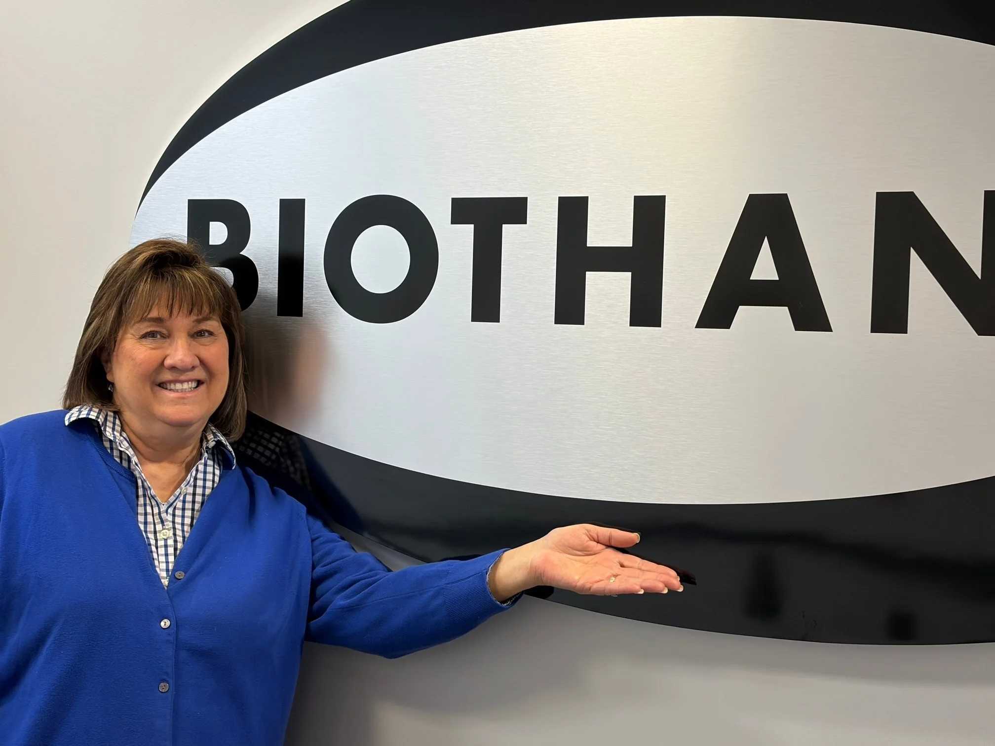A smiling woman in a blue cardigan and checkered shirt standing next to a large sign that says BIOTHAN