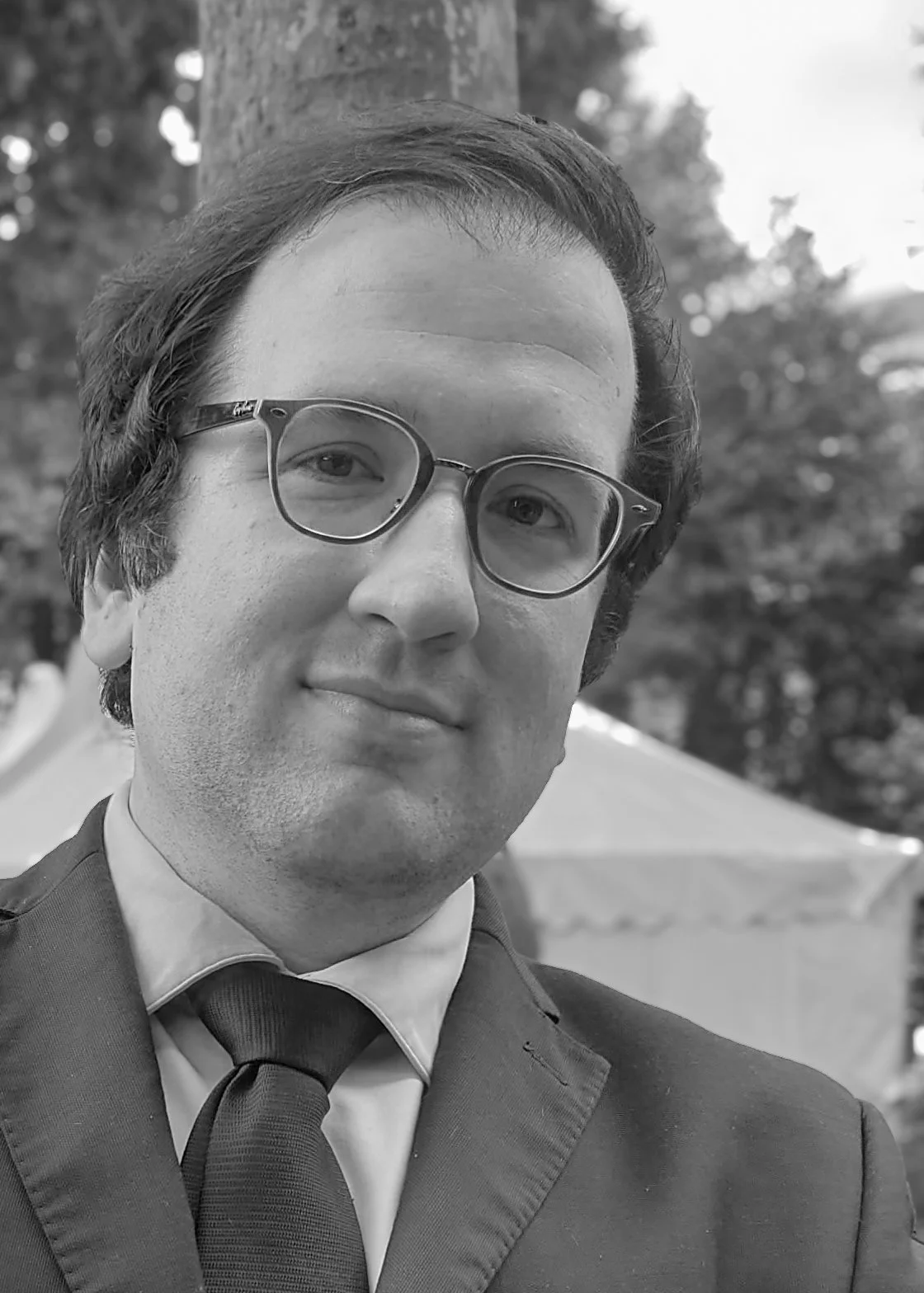 Un uomo con capelli scuri e occhiali, vestito con un abito e una cravatta, posa all'aperto con uno sfondo di alberi e una tenda.