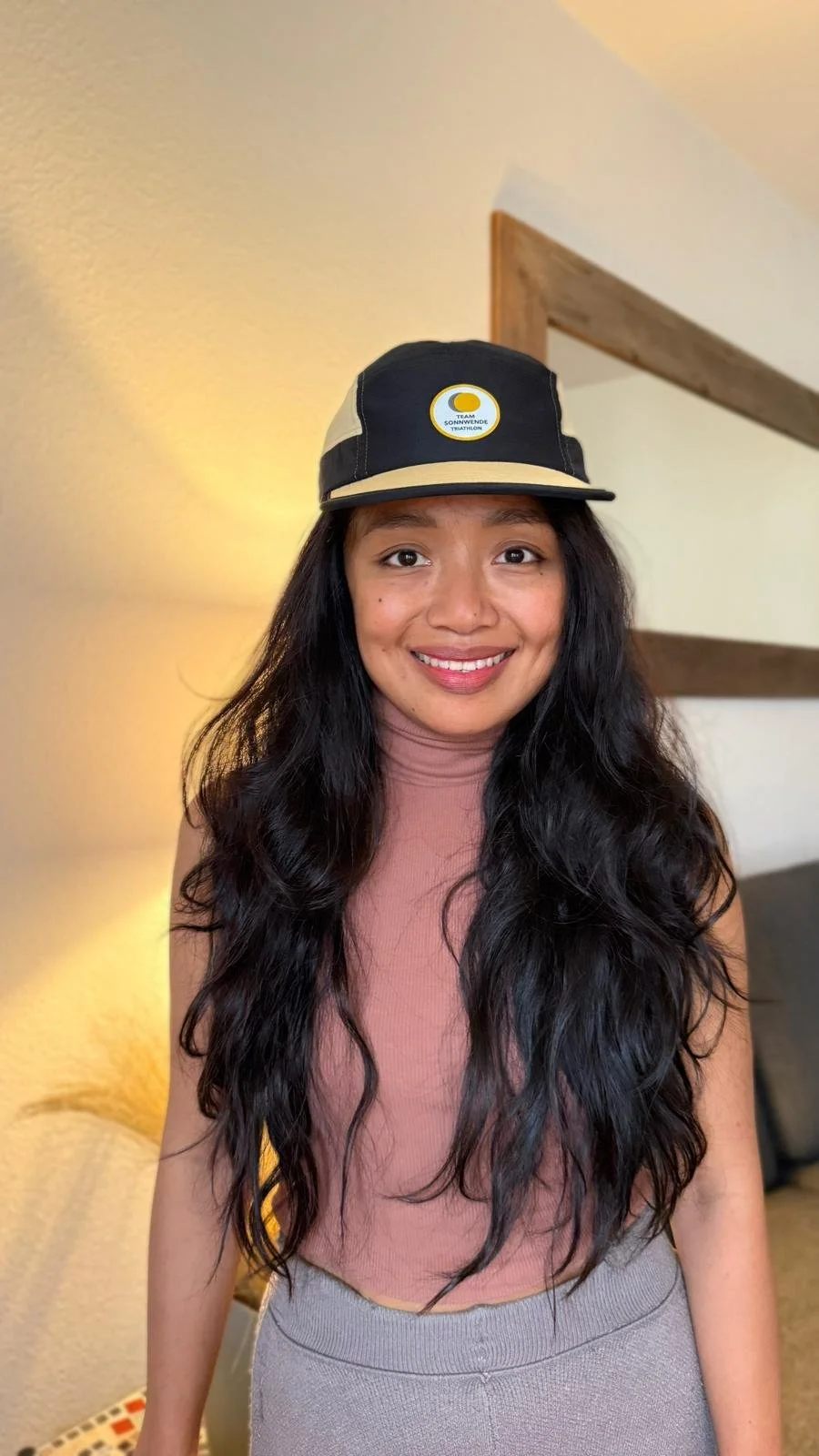 Junge Frau mit langen schwarzen Haaren, trägt eine Kappe mit Sonnenwende-Logo, rosa T-Shirt und graue Hose, lächelnd im Wohnzimmer.