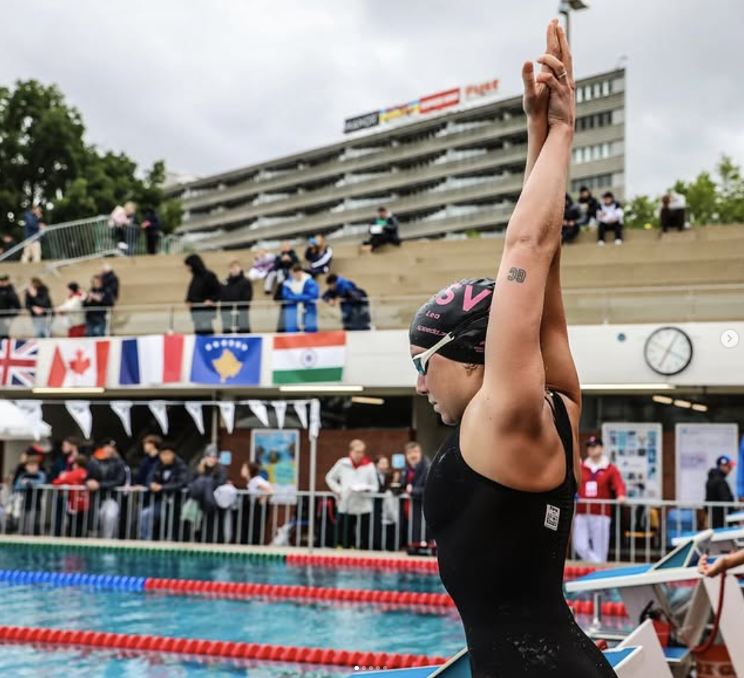 Schwimmerin bereitet sich am Beckenrand vor, während Zuschauer und Teammitglieder im Hintergrund sitzen und internationales Flaggendesign sichtbar ist.