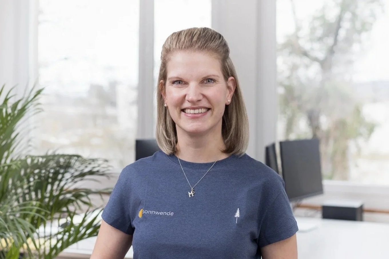 Lächelnde junge Frau in einem Büro, trägt ein dunkelblaues T-Shirt mit Sonnenwende-Logo und einem Anhänger mit Initialen. Hinter ihr sind große Fenster und Büromöbel.