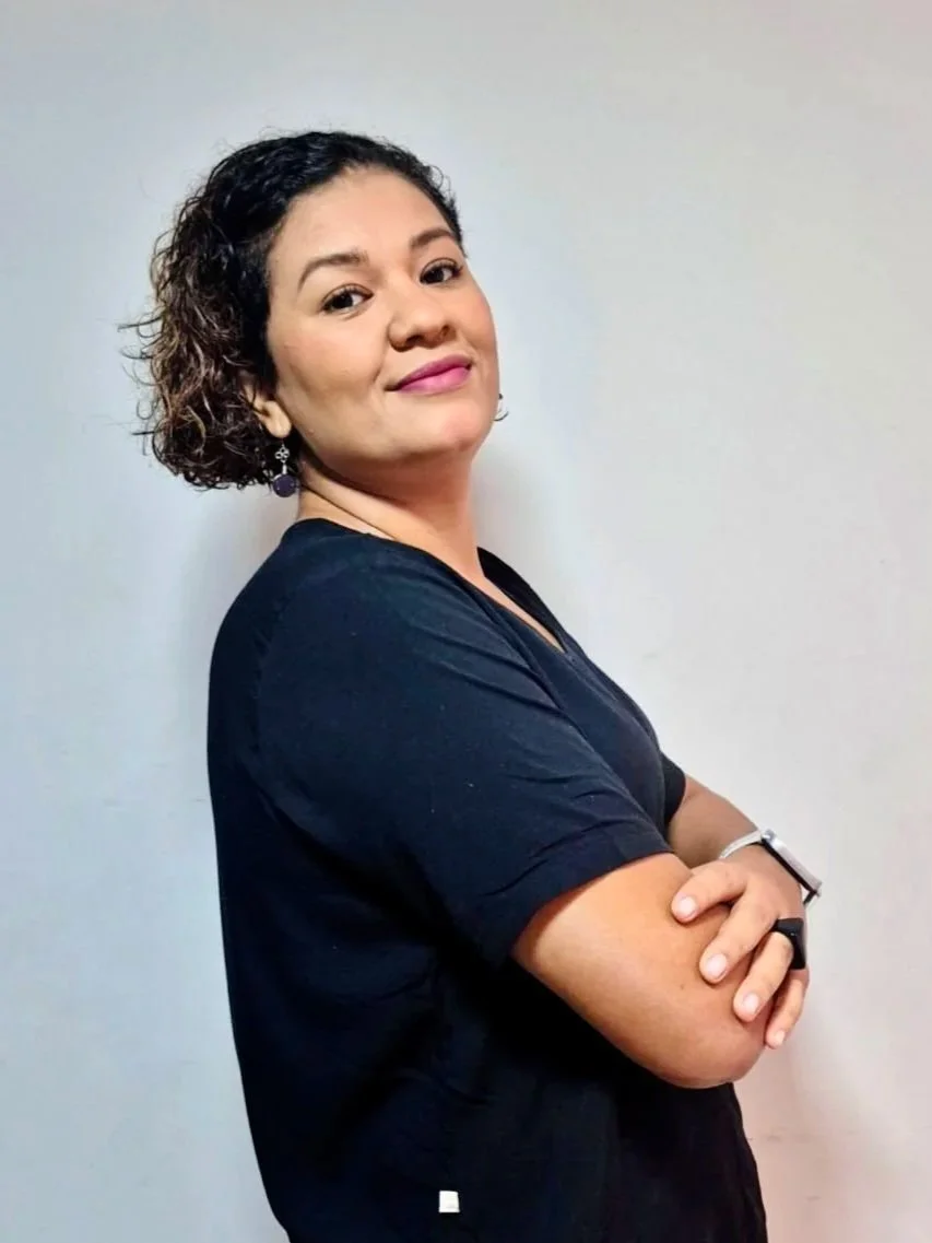 Mulher com cabelos curtos e cacheados, posando com os braços cruzados, vestindo camiseta preta, com fundo branco.