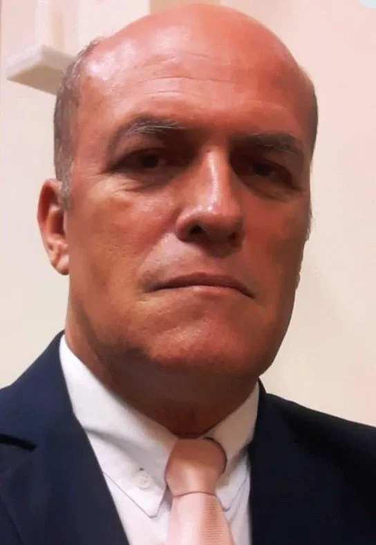 Homem de terno escuro, camisa branca e gravata rosa, com expressão séria, em um fundo claro.