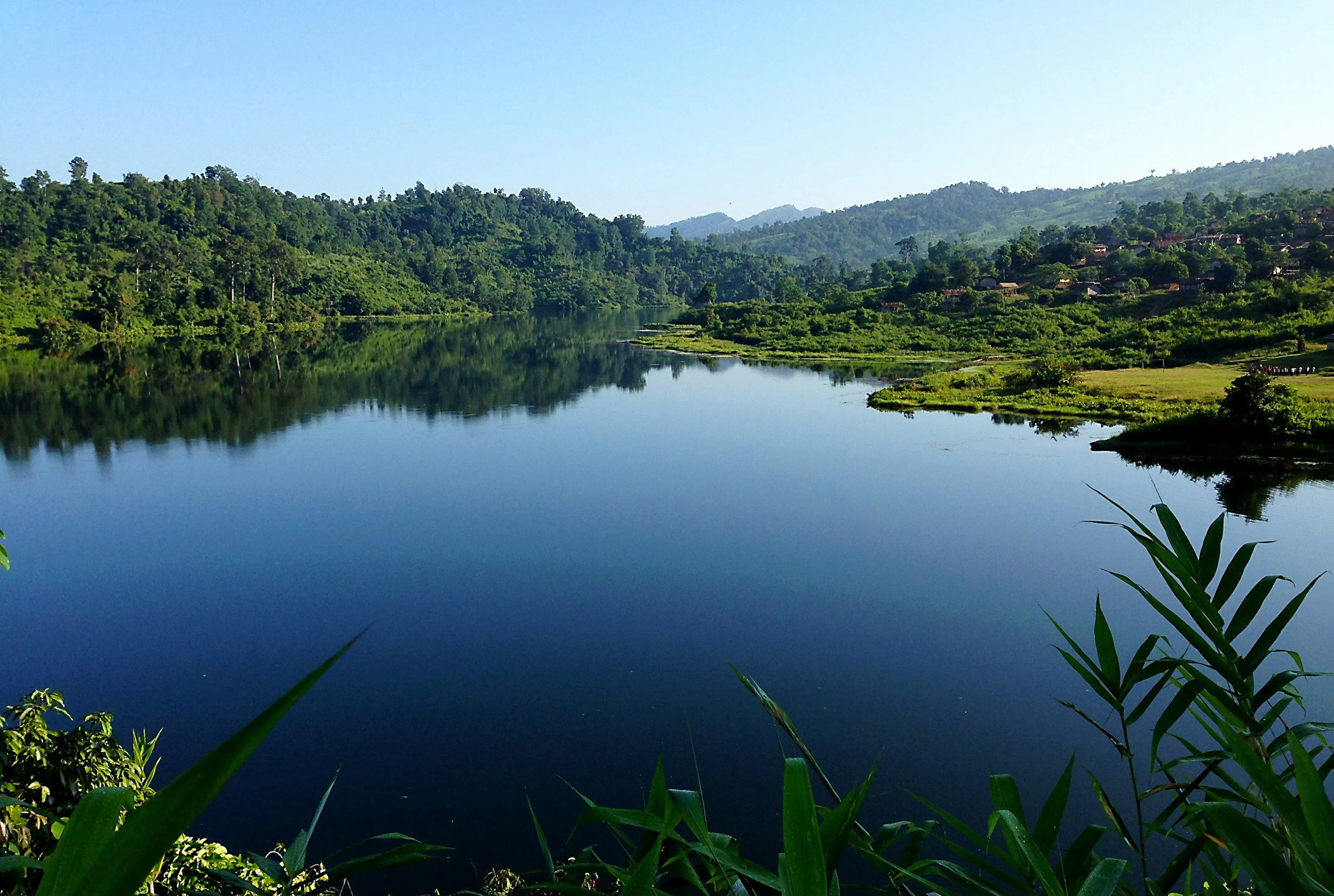 Calm river flowing through lush green landscape with hills in the background, under a clear blue sky.