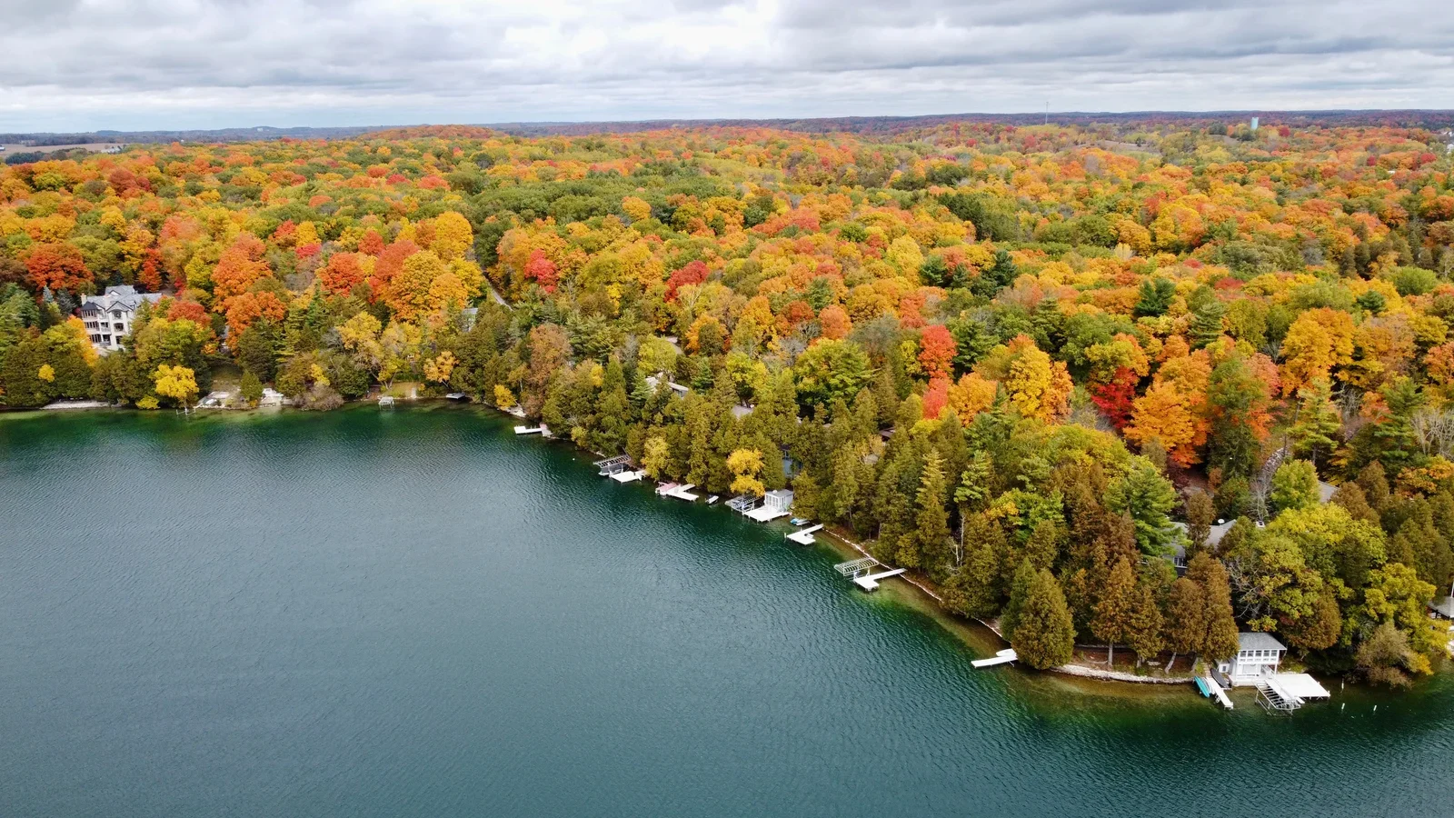 Aerial view of a lake bordered by a dense forest with trees in vibrant autumn colors. Several houses with docks line the shoreline.
