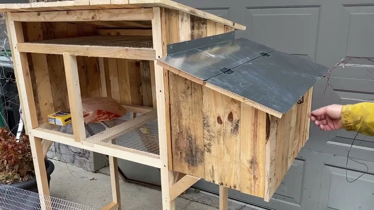 A wooden chicken coop with one open side and a slanted metal roof, situated outdoors.