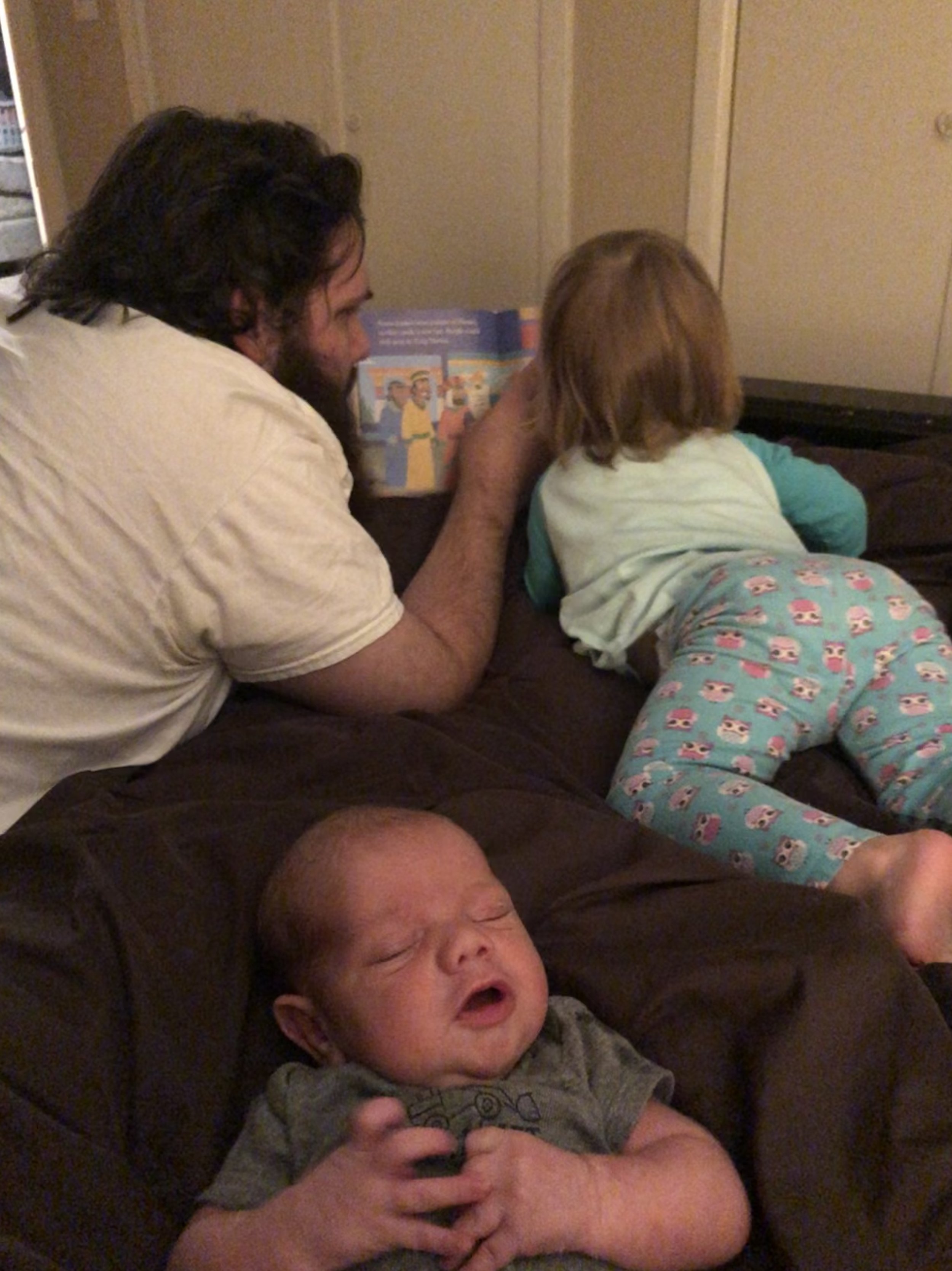 A man with long dark hair and a beard is going down on a woman leaning over a black piece of furniture, possibly a bed, while a young child with a bald head is sleeping on the same piece of furniture in front of them.