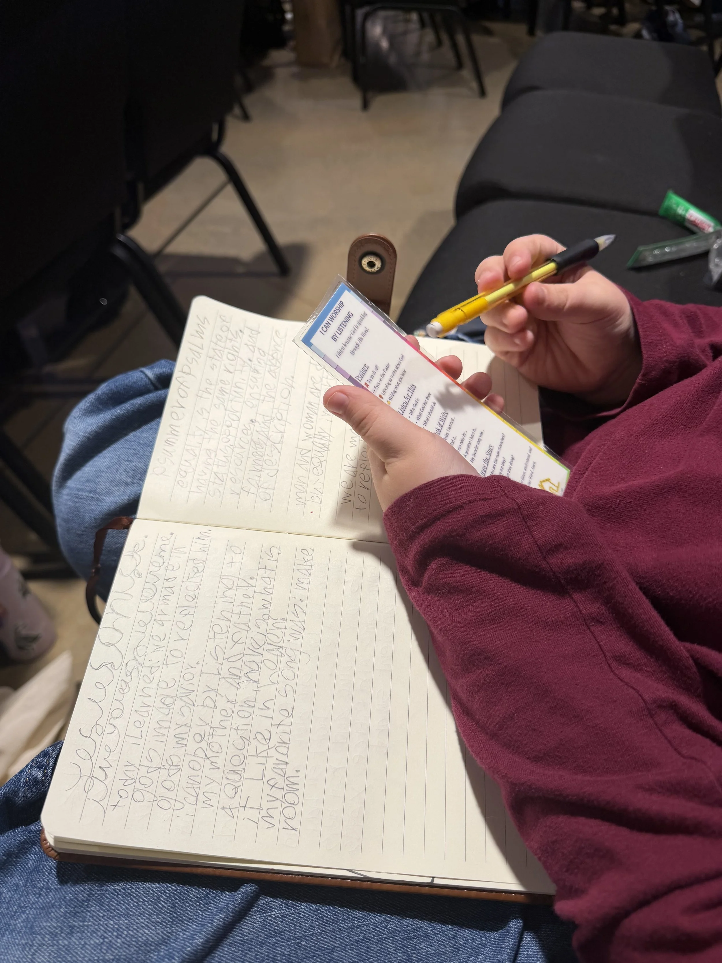 A person wearing a red long sleeve shirt is sitting and writing in a notebook with lined paper. Another hand is holding a yellow and black pen. The notebook has handwritten text. On the person's lap are blue jeans. There are some objects on the right side of the seat, such as a green tube and a plastic bag. The background shows several black chairs and a wooden floor.