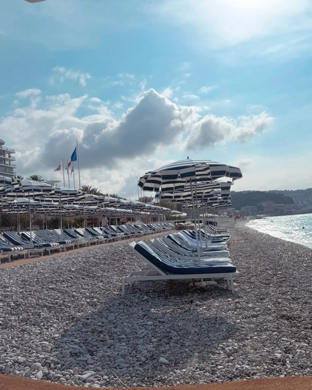 Empty beach with striped lounge chairs and umbrellas, with flags in the background and a cloudy sky.