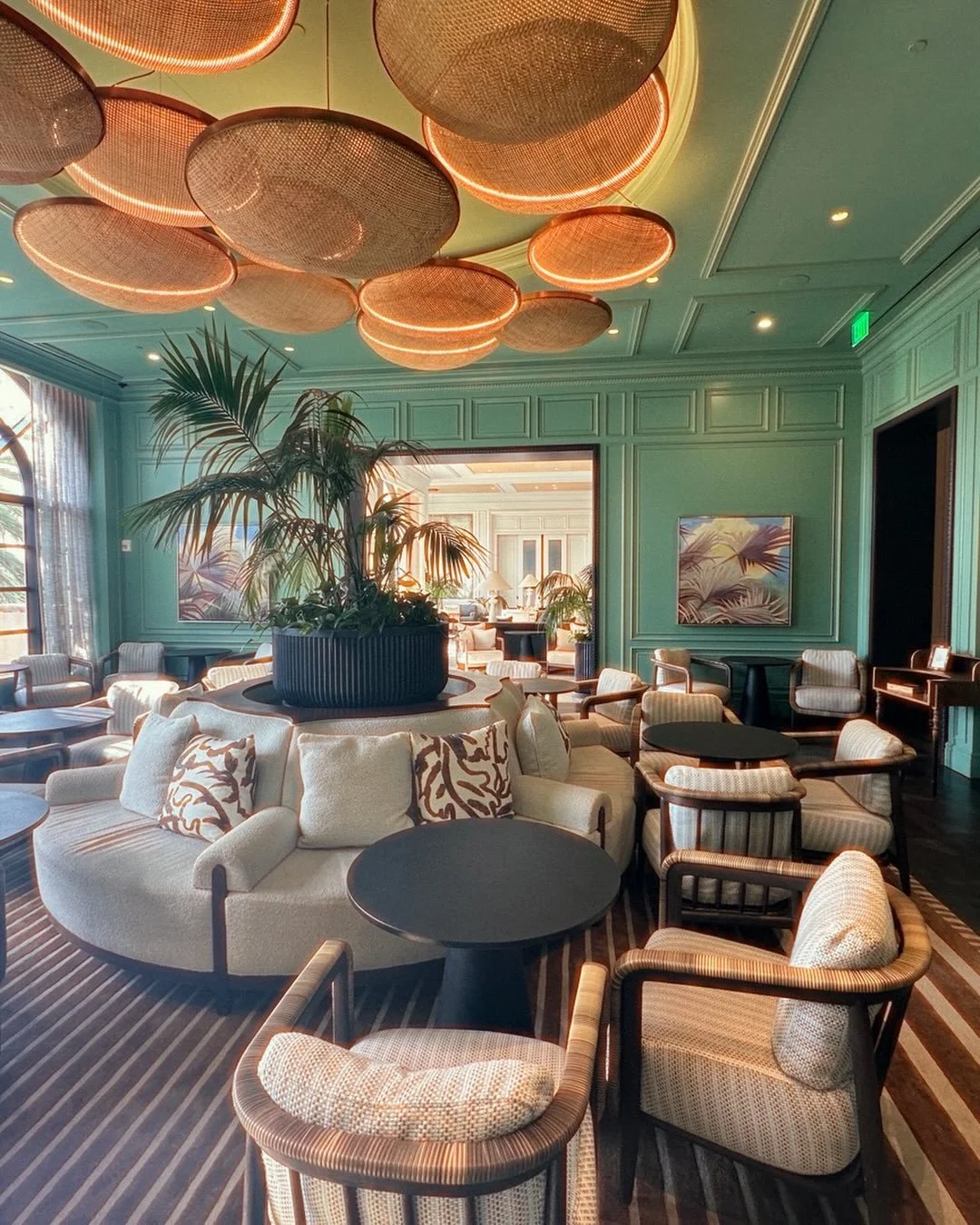 Elegant hotel lounge with green paneled walls, circular lighting fixtures on the ceiling, a large central plant, and various seating options including a sectional sofa with decorative pillows and armchairs around small tables.