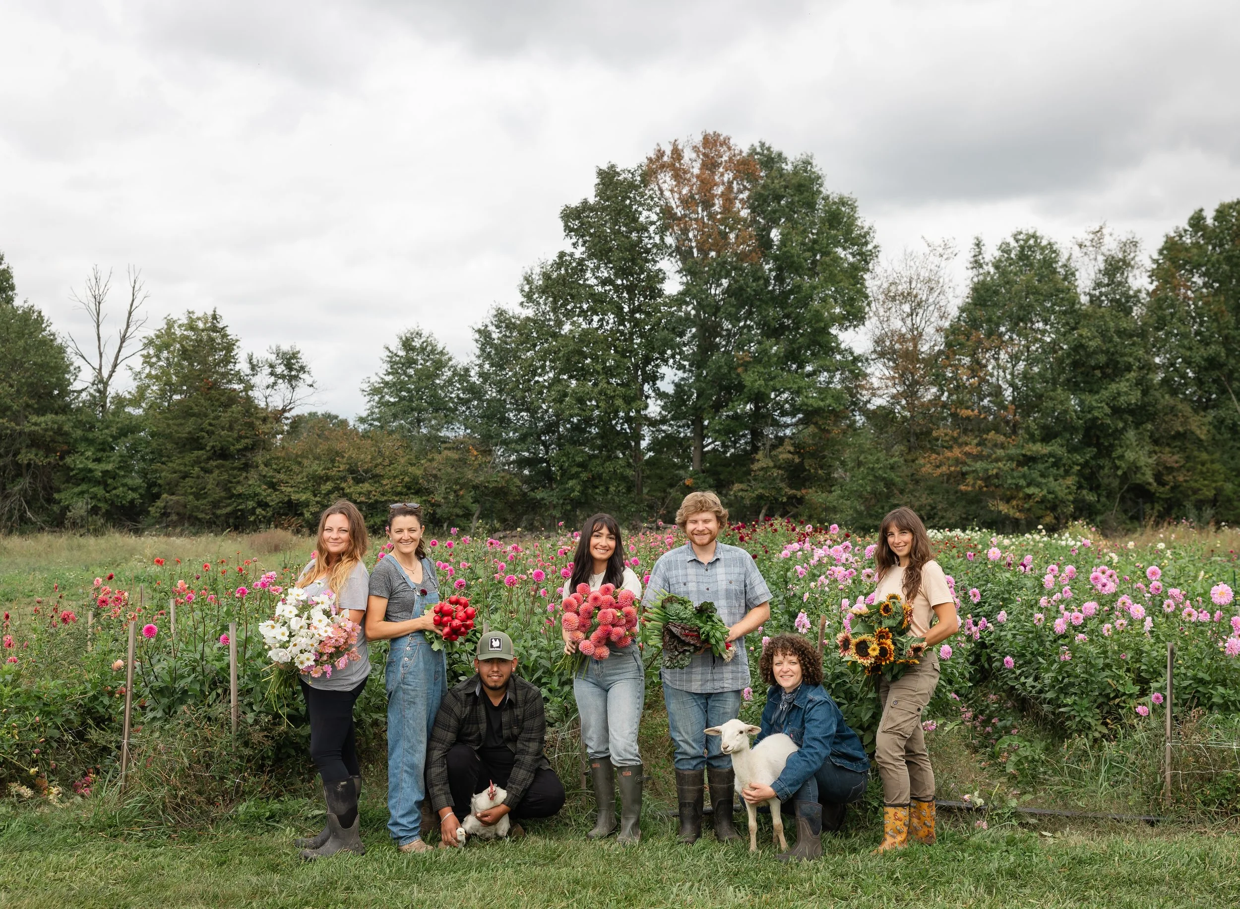 A group of people standing in a farm field surrounded by flowers, with trees in the background and cloudy sky above.