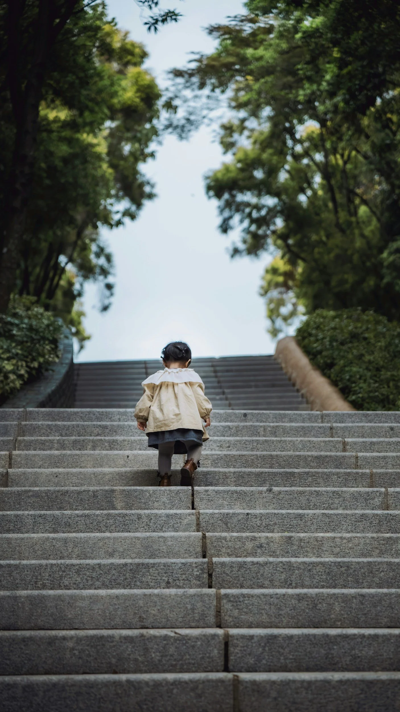 Toddler in a white dress walks up concrete steps. At the top of the steps, trees and a light blue sky are visible.